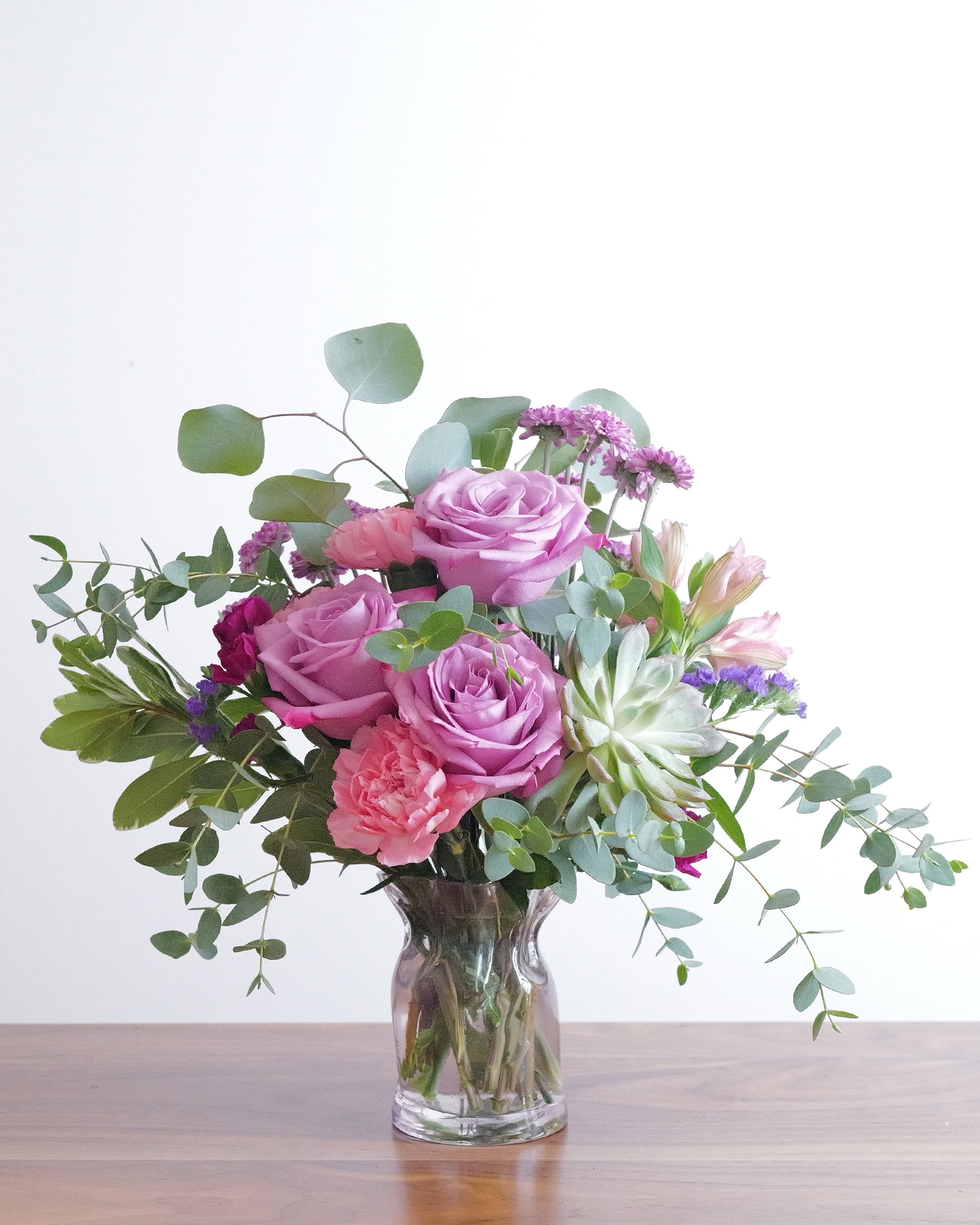 Lavender roses, pink carnations and an echeveria succulent with eucalyptus in a glass vase on a wooden table