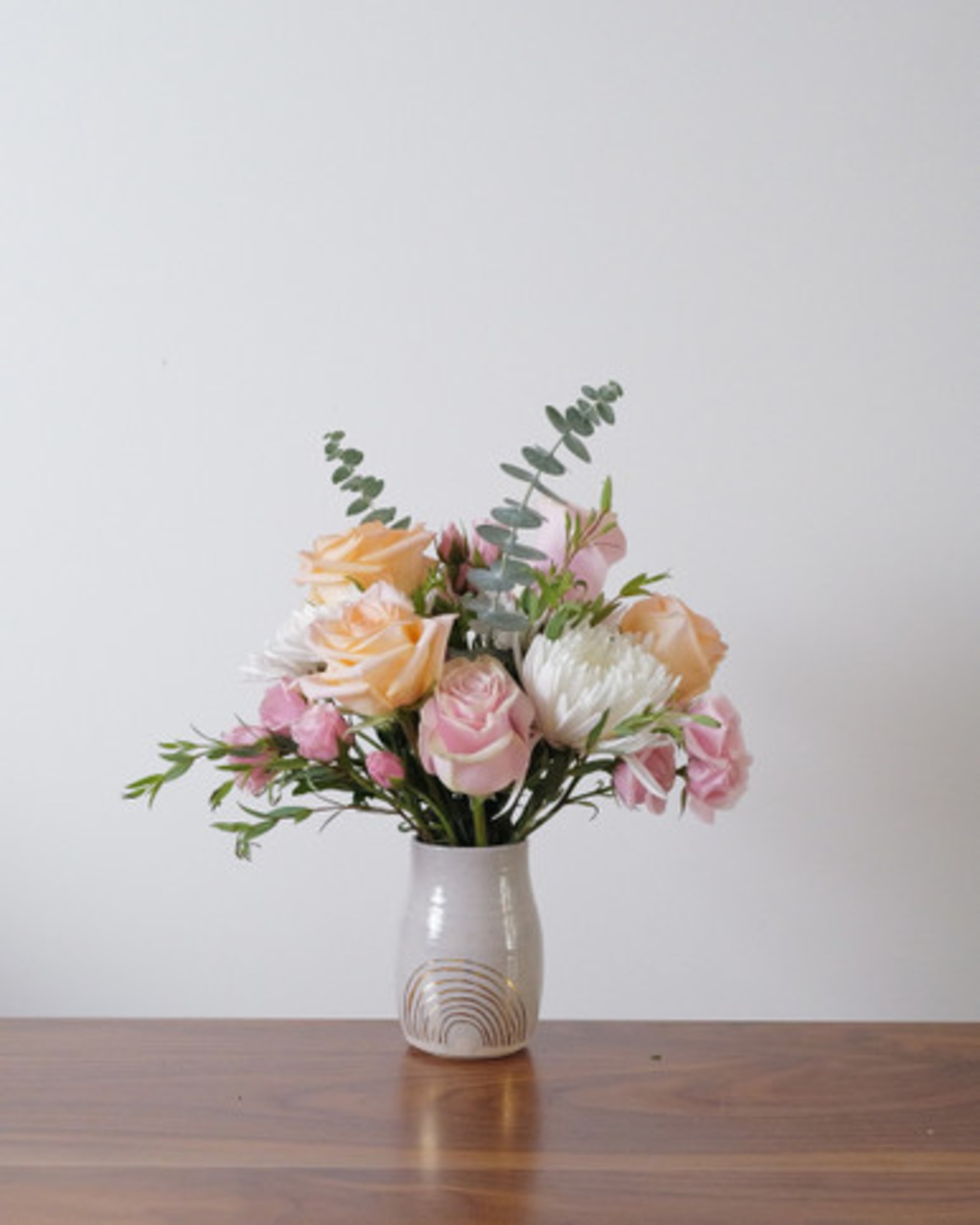 Peach and pink roses with white chrysanthemums and eucalyptus in a white ceramic vase with a gold rainbow, on a wood table