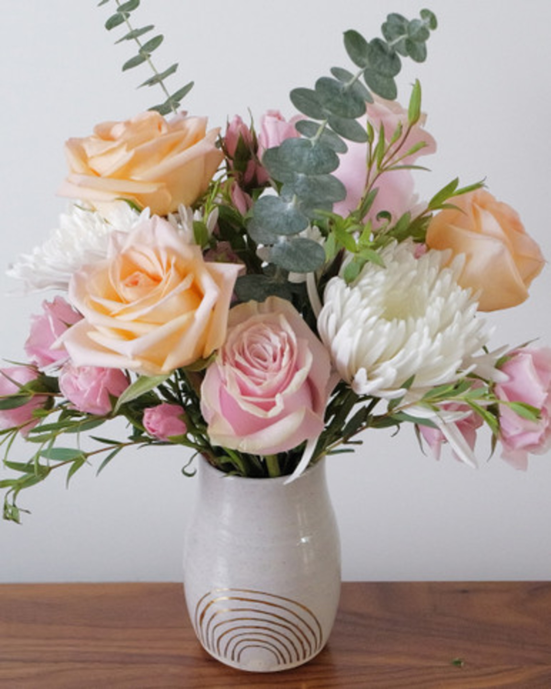 Close view of peach and blush roses, white chrysanthemums and silvery eucalyptus in a white vase painted with a gold rainbow