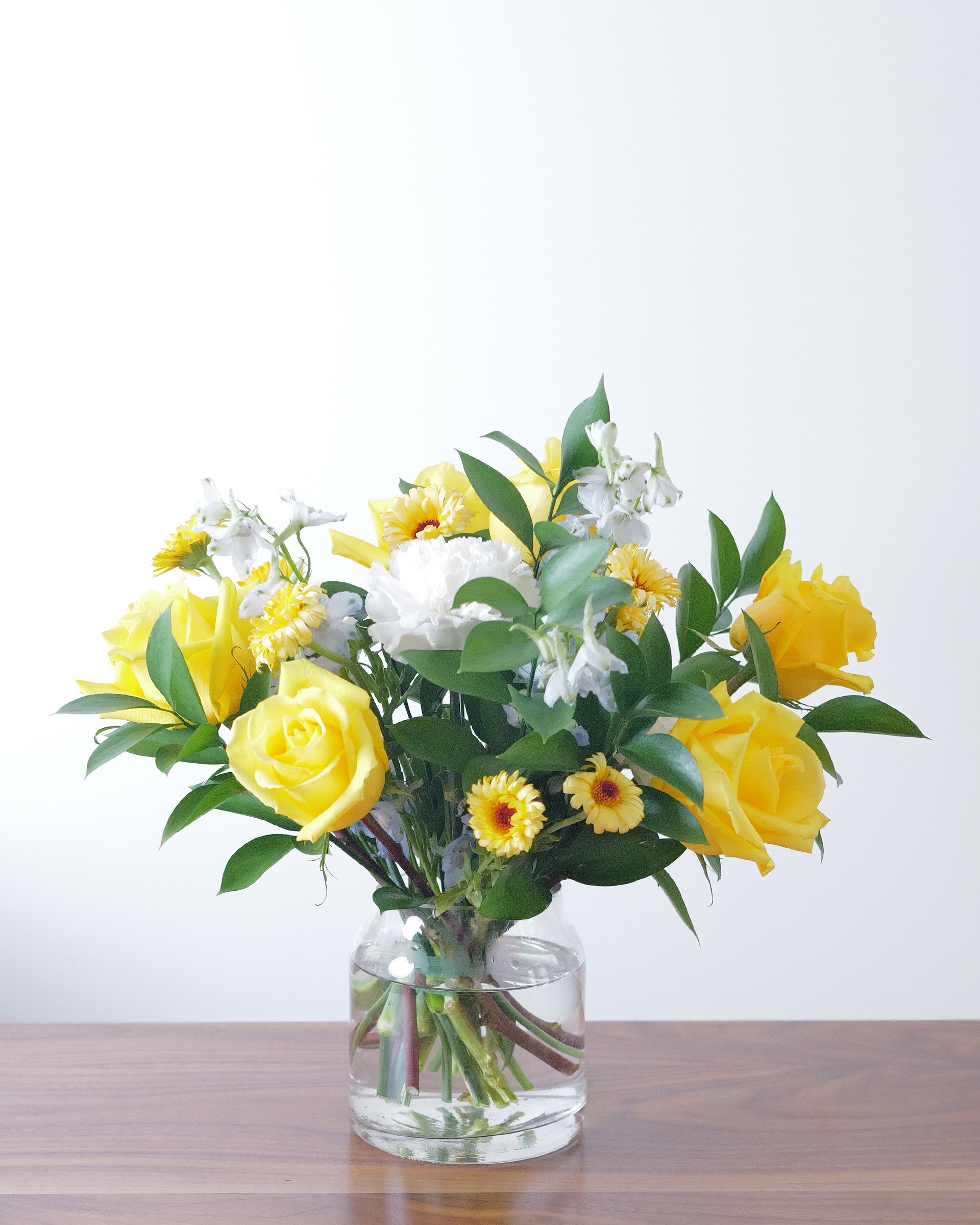 Yellow roses, golden daisy-like blooms, a white carnation and pale blue-white flowers with greens in a clear glass vase on