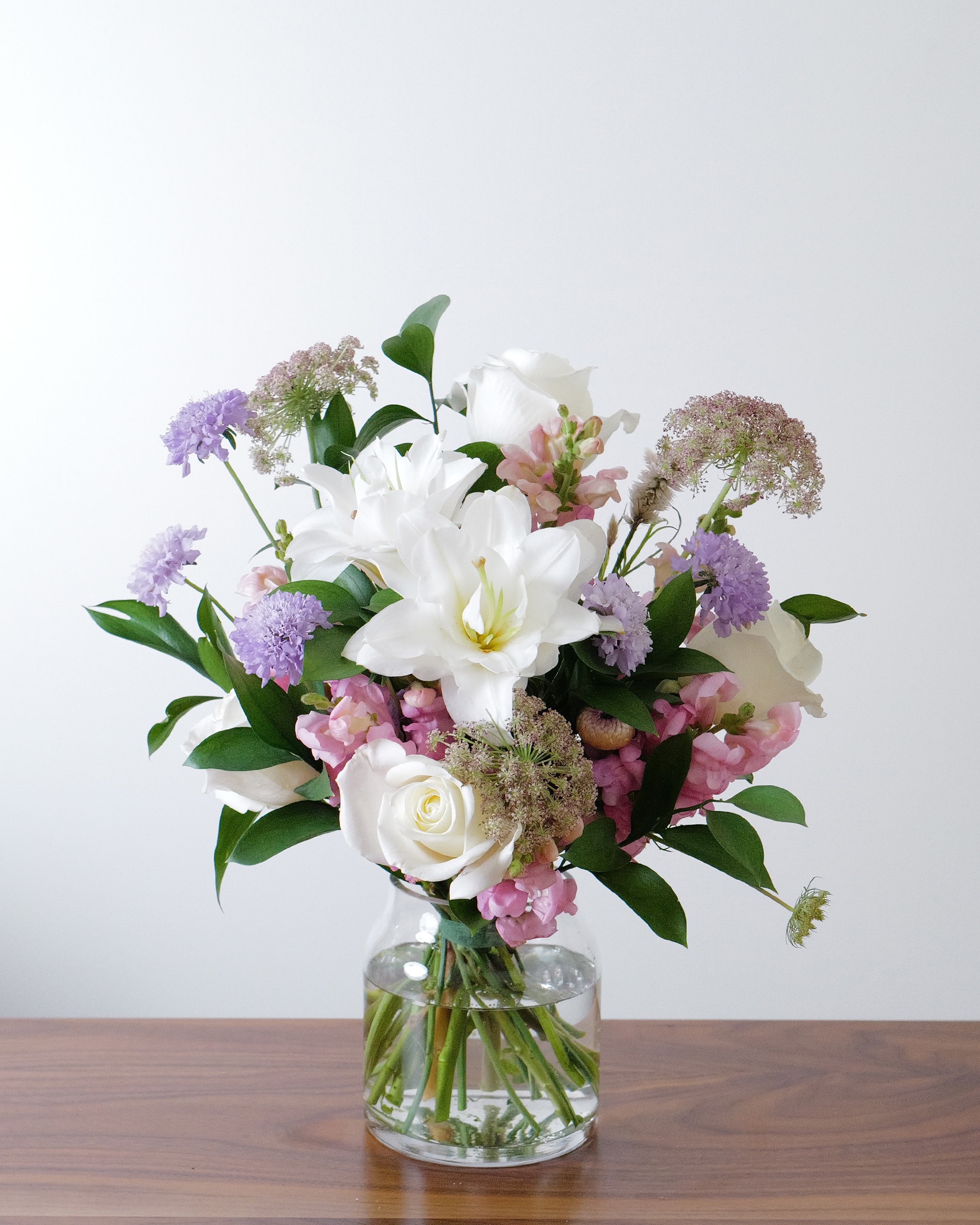 White lilies, white roses, pink snapdragons and lavender blooms with greens in a clear glass vase on a wooden table