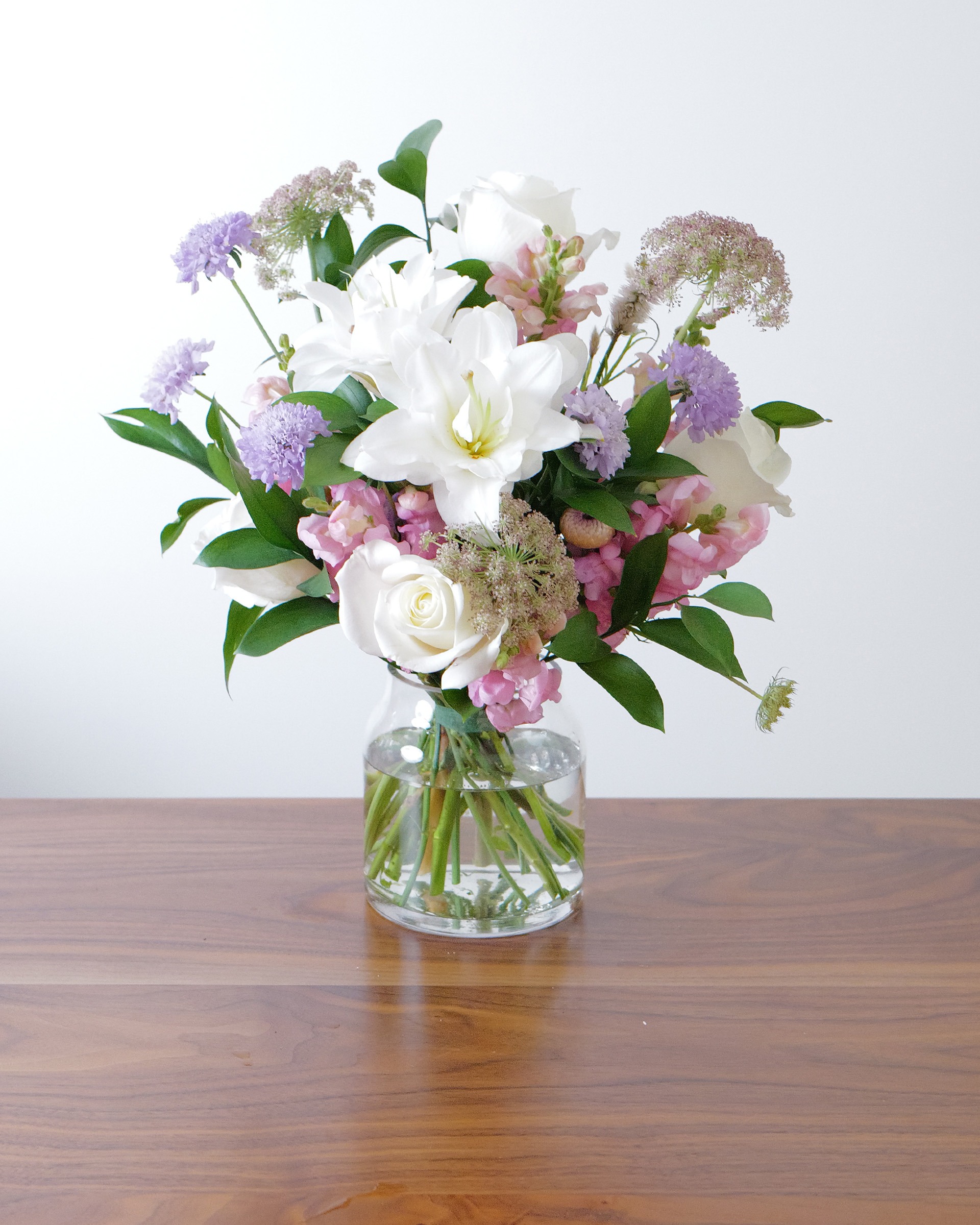 Soft pink, lavender and white garden-style bouquet in a clear glass vase on a walnut table
