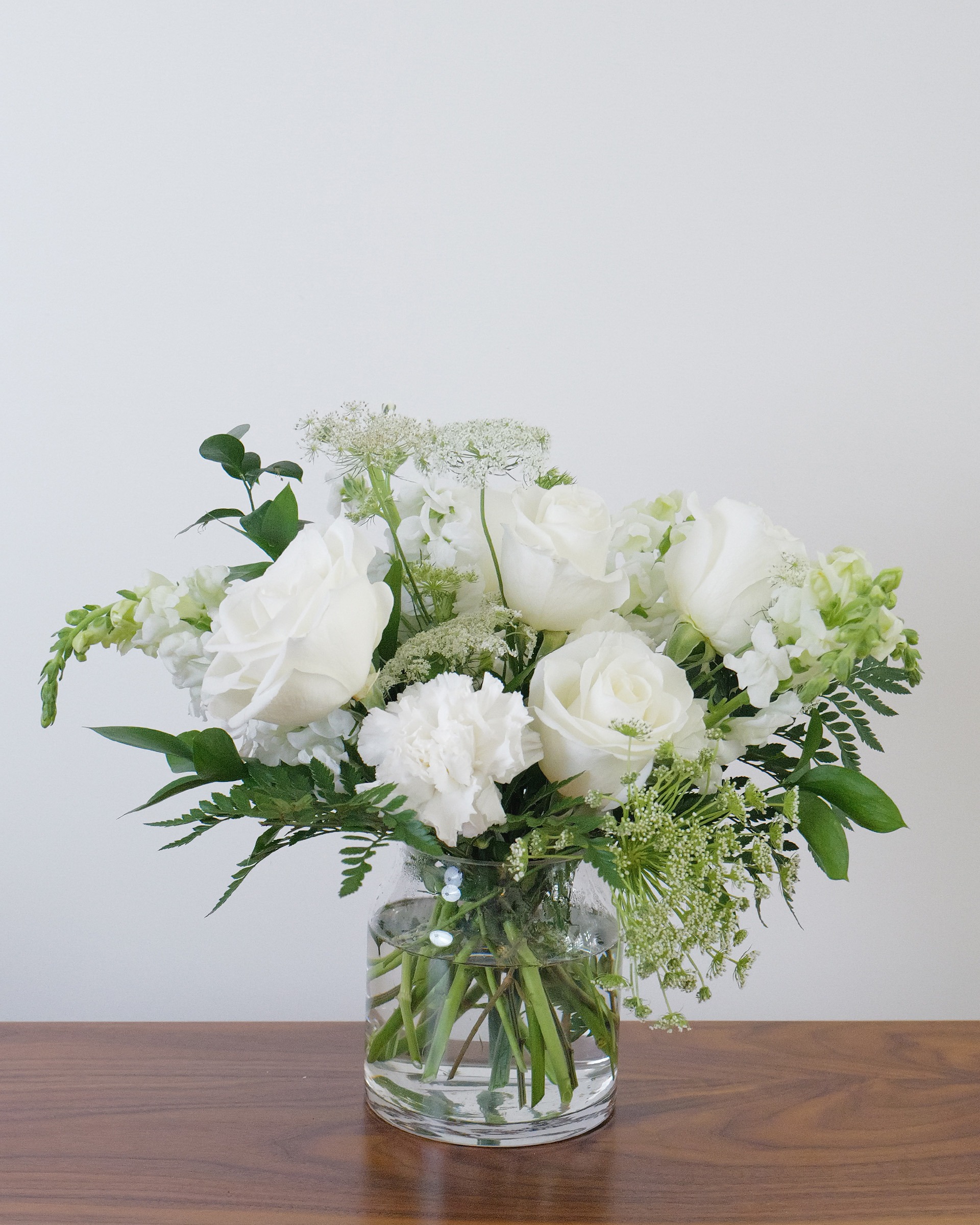 White roses, carnation, snapdragons and lacy white blooms with greens in a clear glass vase on a wood table