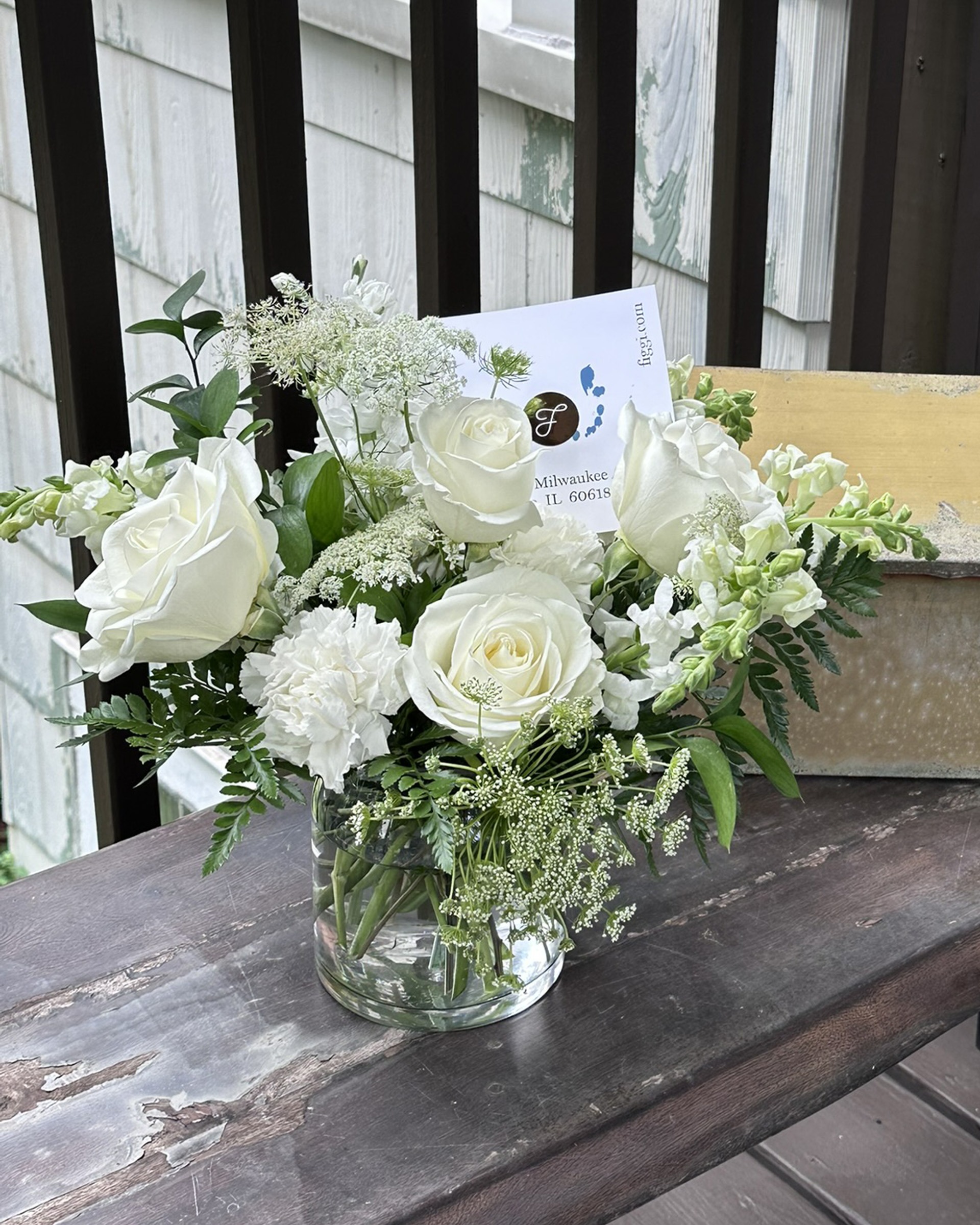 All-white arrangement of roses and snapdragons in a clear glass vase with a Figgi card, on a weathered porch rail