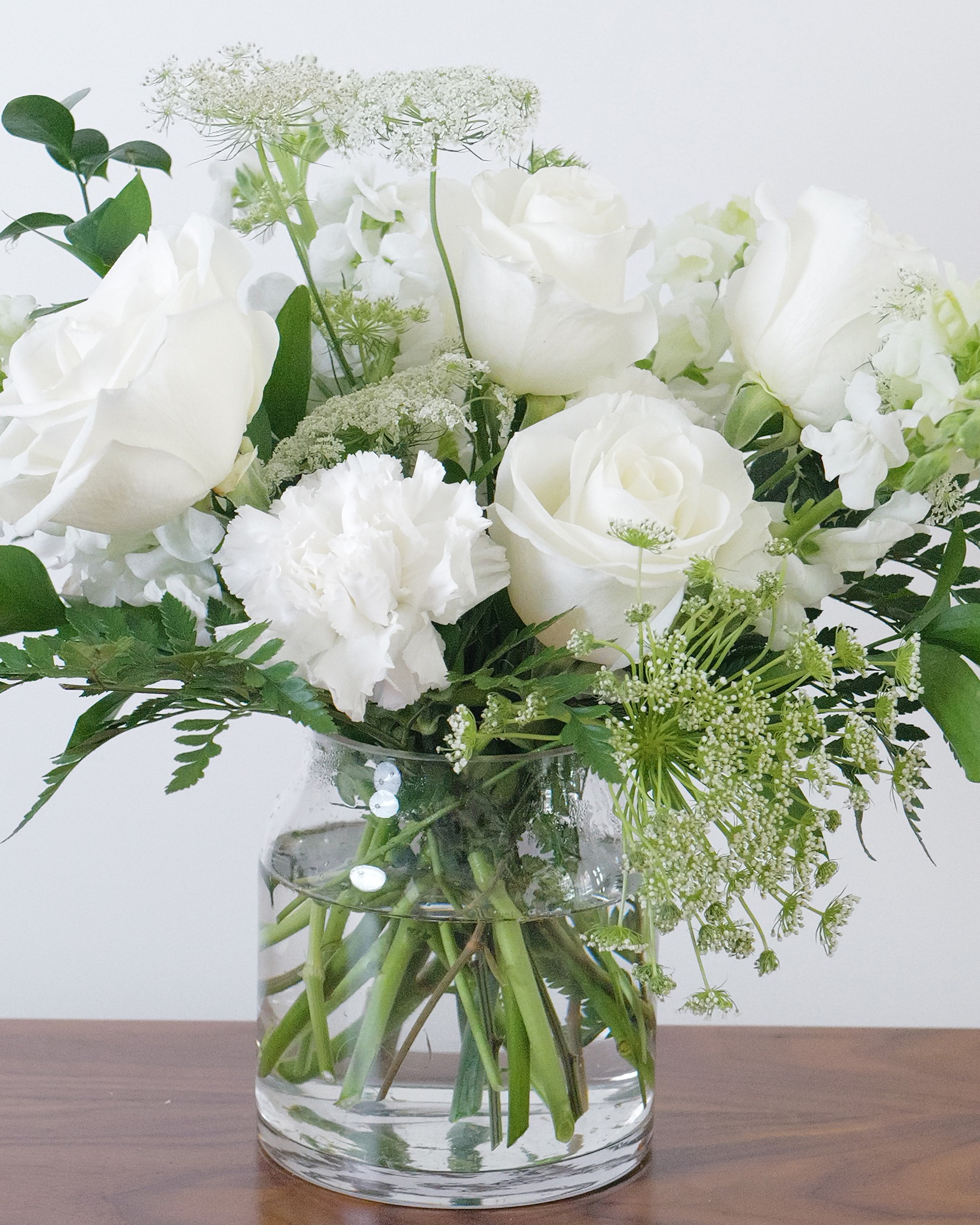 Close view of white roses, a white carnation and lacy white blooms with ferns in a clear glass vase