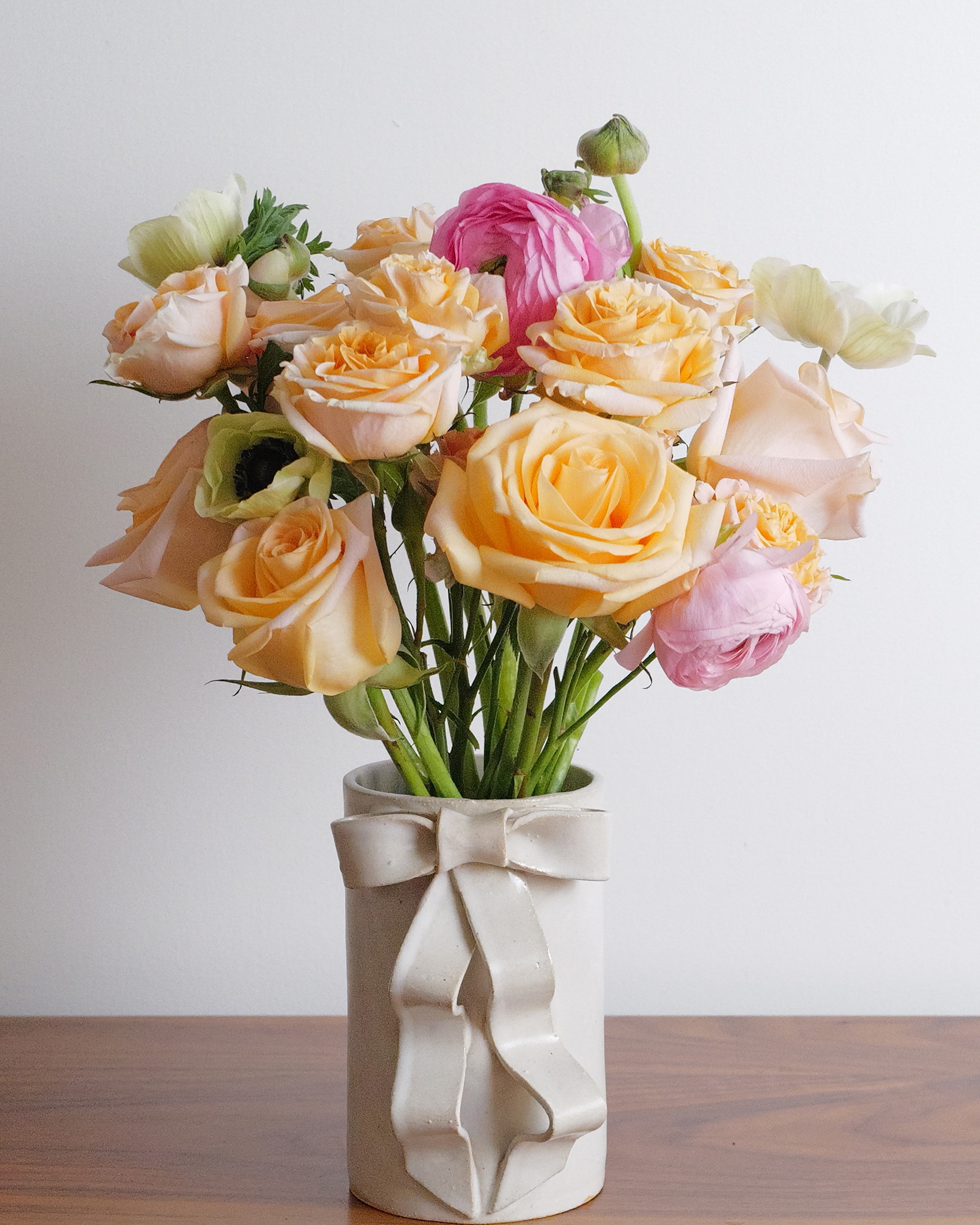 Peach roses, pink ranunculus and pale anemones in a cream ceramic vase with a sculpted bow, on a wooden table