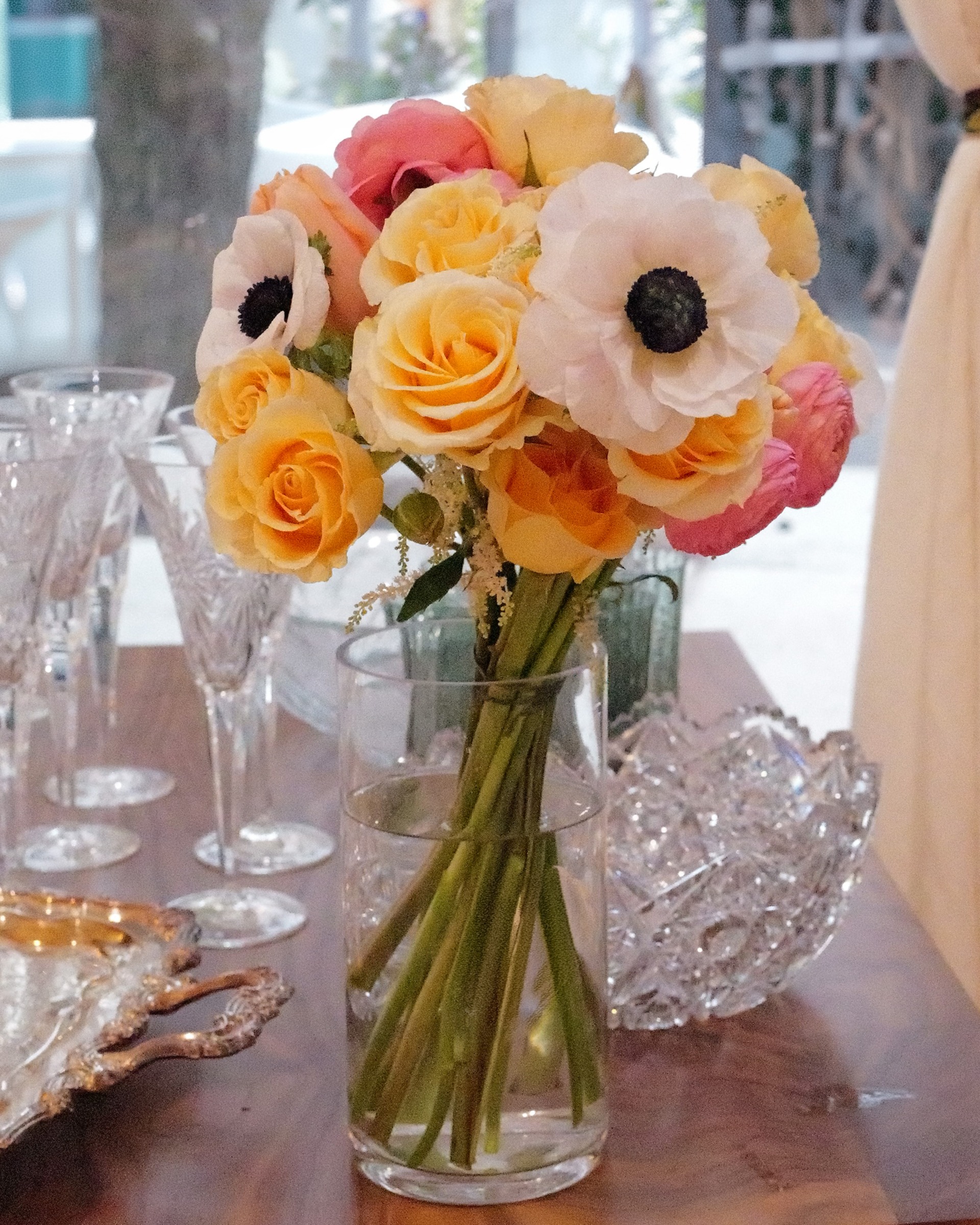 Peach roses, pink ranunculus and white anemones in a clear glass vase beside crystal glasses and a silver tray