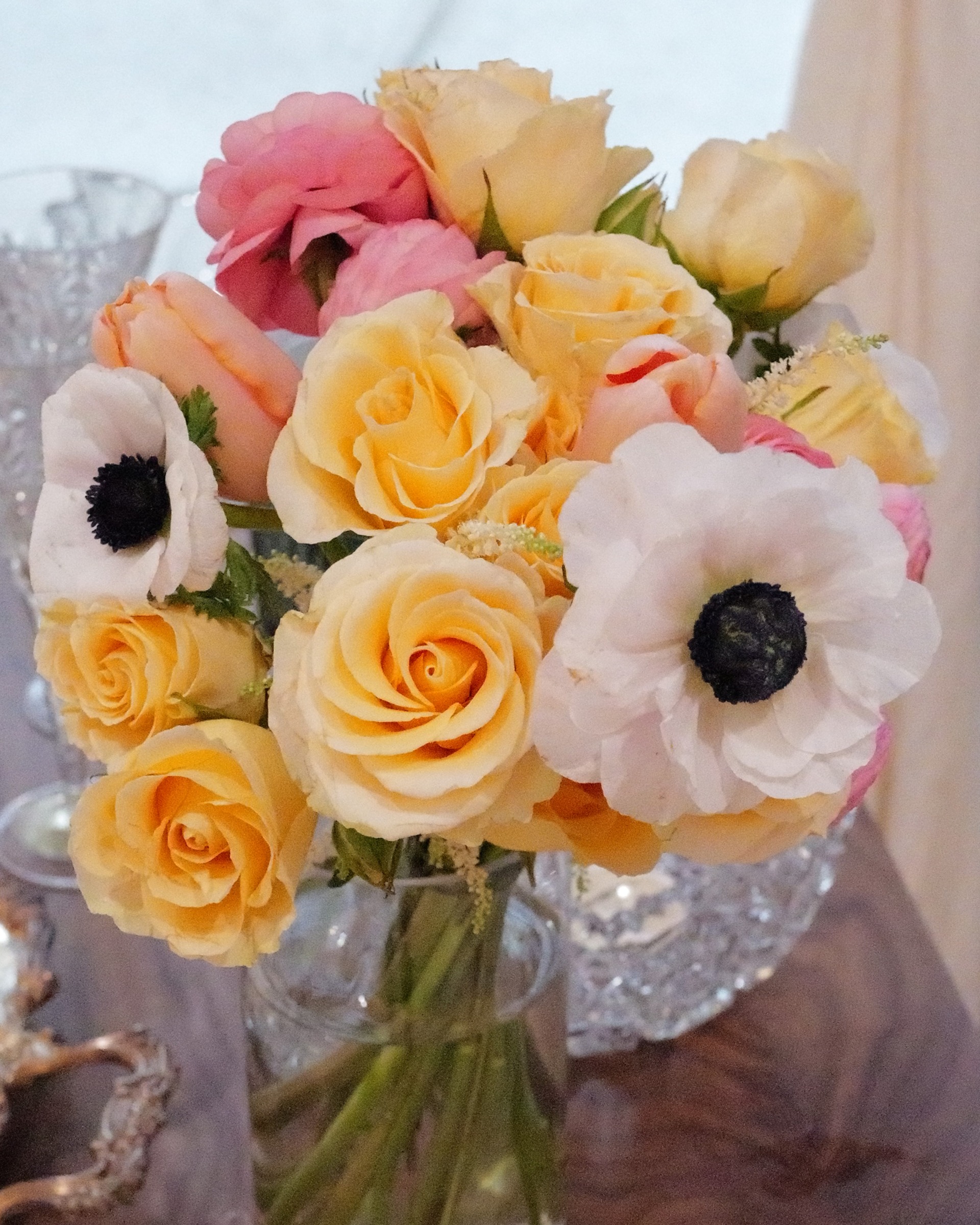 Close view of peach roses, pink ranunculus and white anemones with dark centers above cut crystal on a wooden table