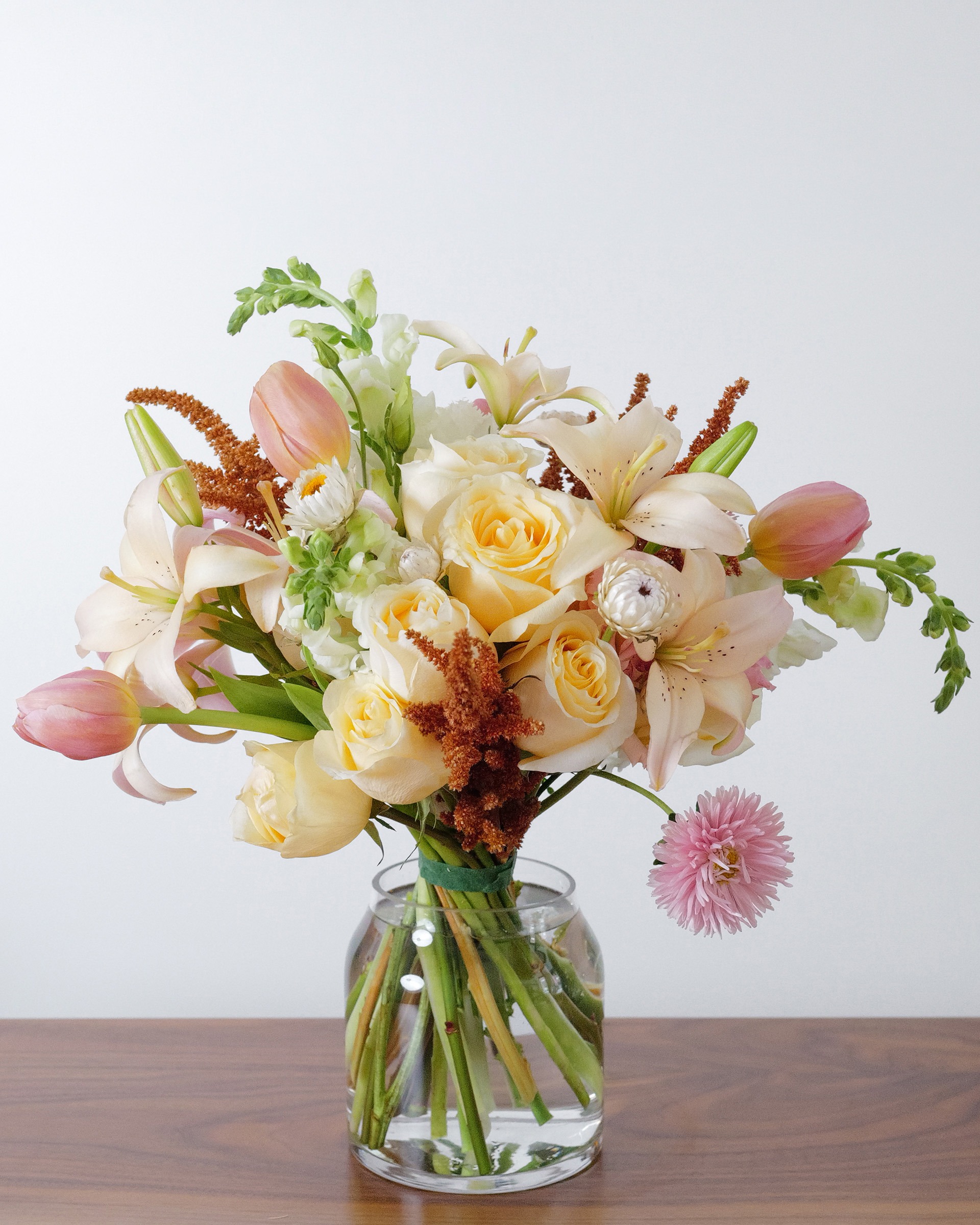 Peach roses, blush lilies and pink tulips with rust-orange accents in a clear glass vase on a wooden table
