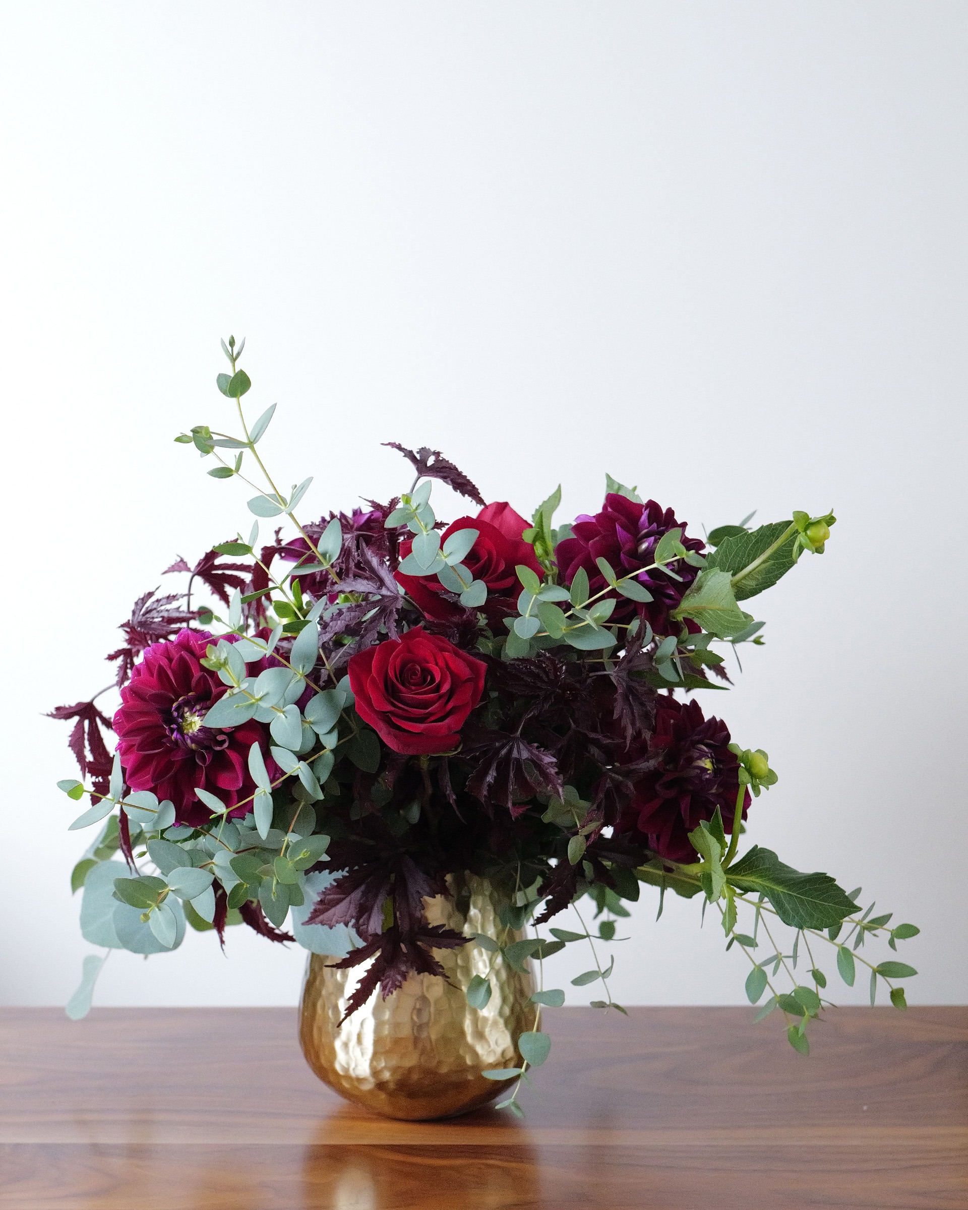 Deep red roses and burgundy dahlias with eucalyptus and dark foliage in a hammered gold vase on a wooden table