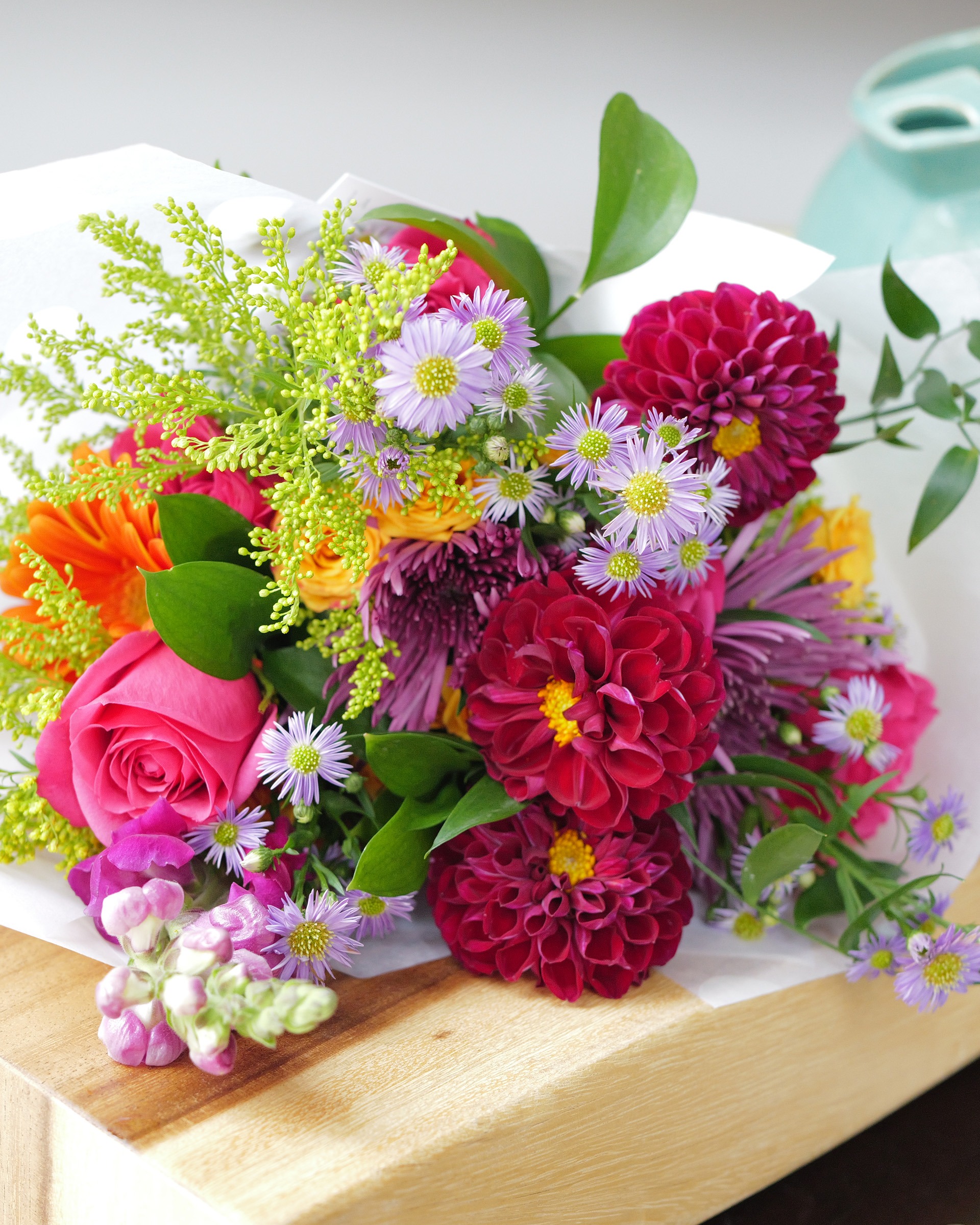 Bright wrapped bouquet of magenta dahlias, pink roses, orange gerbera and lavender asters on a wooden board