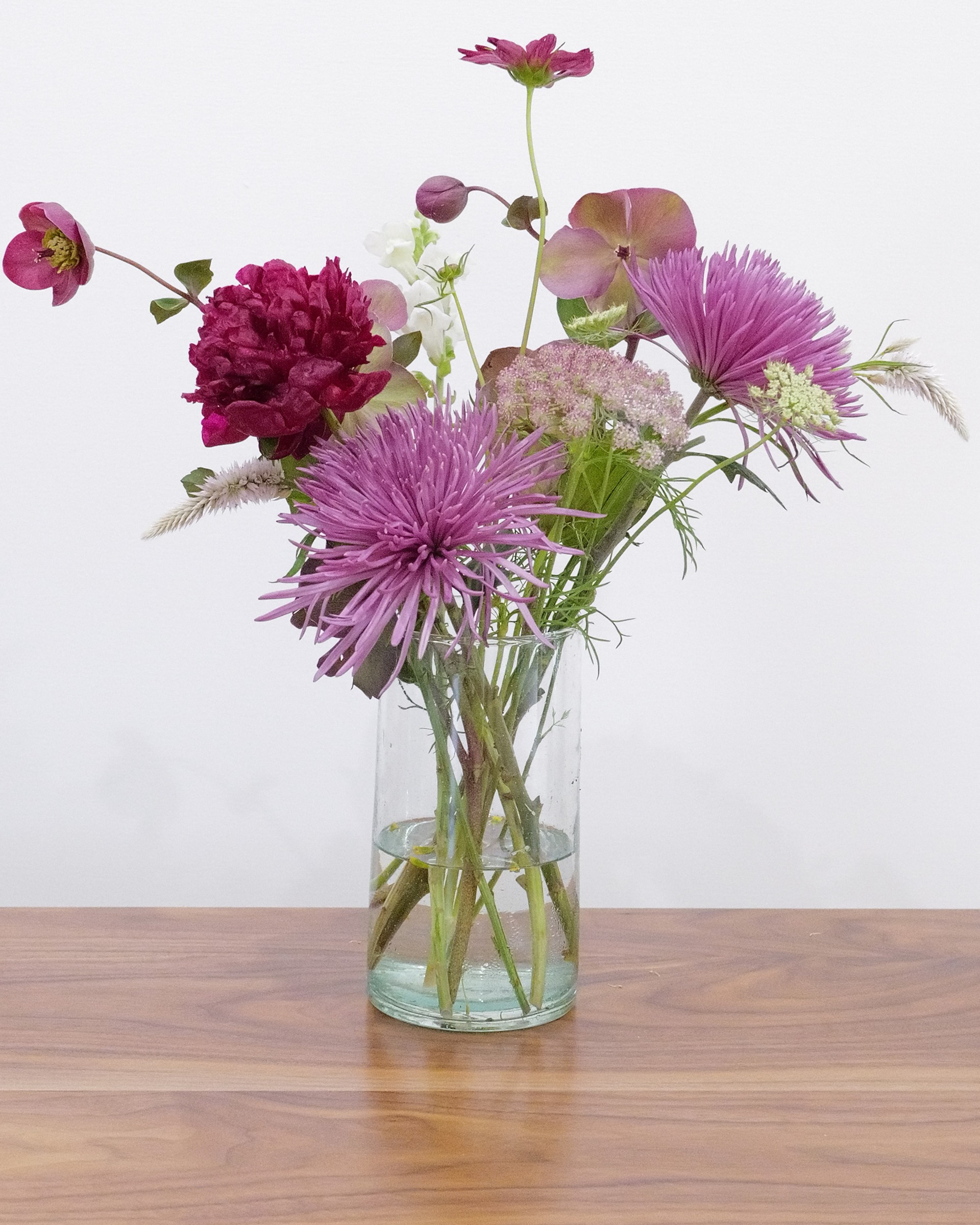 Airy arrangement of purple spider mums, a crimson peony and hellebores in a tall clear glass cylinder on wood