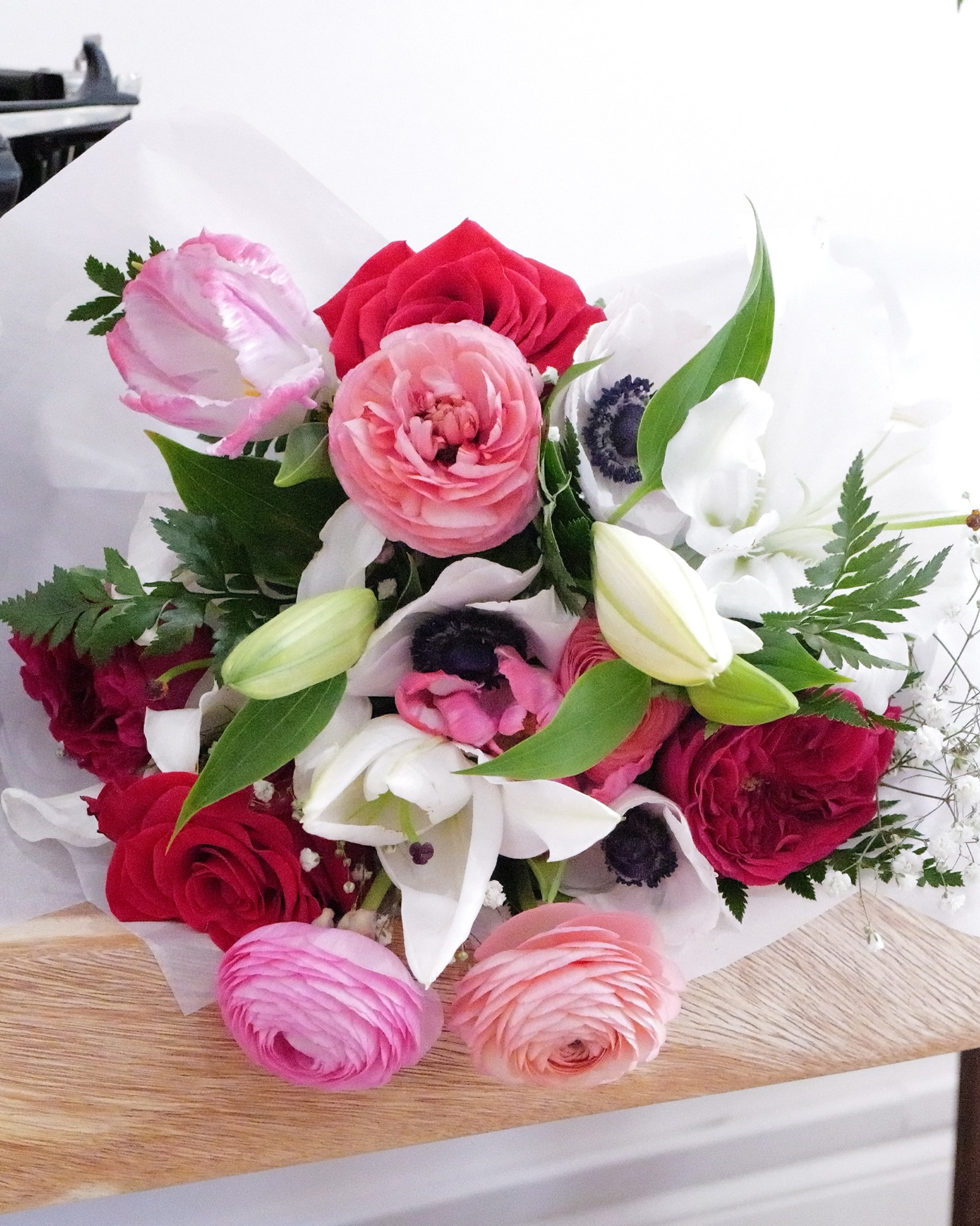 Wrapped bouquet of red and pink roses, pink ranunculus, white anemones and white lilies on a wooden surface