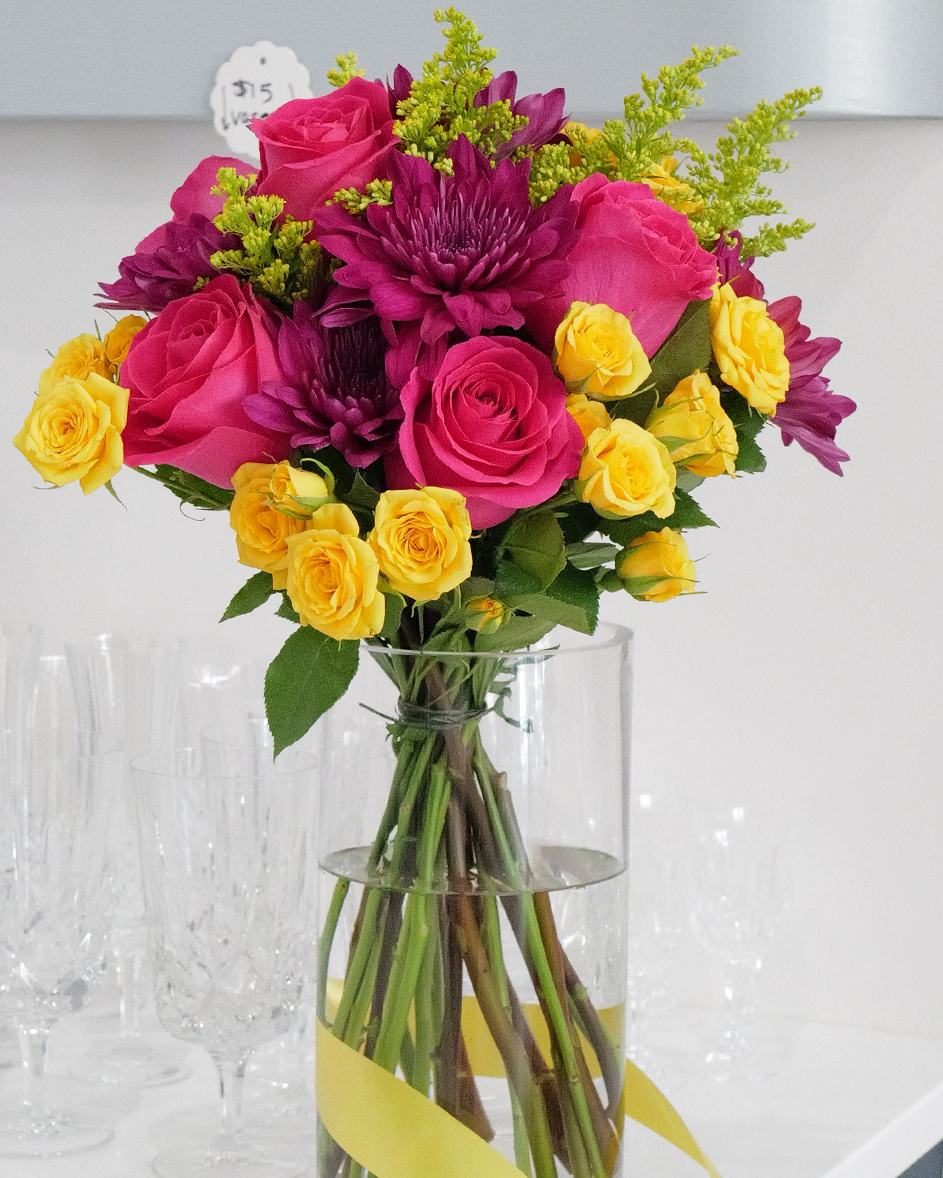 Hot pink roses, purple mums and yellow spray roses in a clear glass vase tied with yellow ribbon, crystal glasses behind