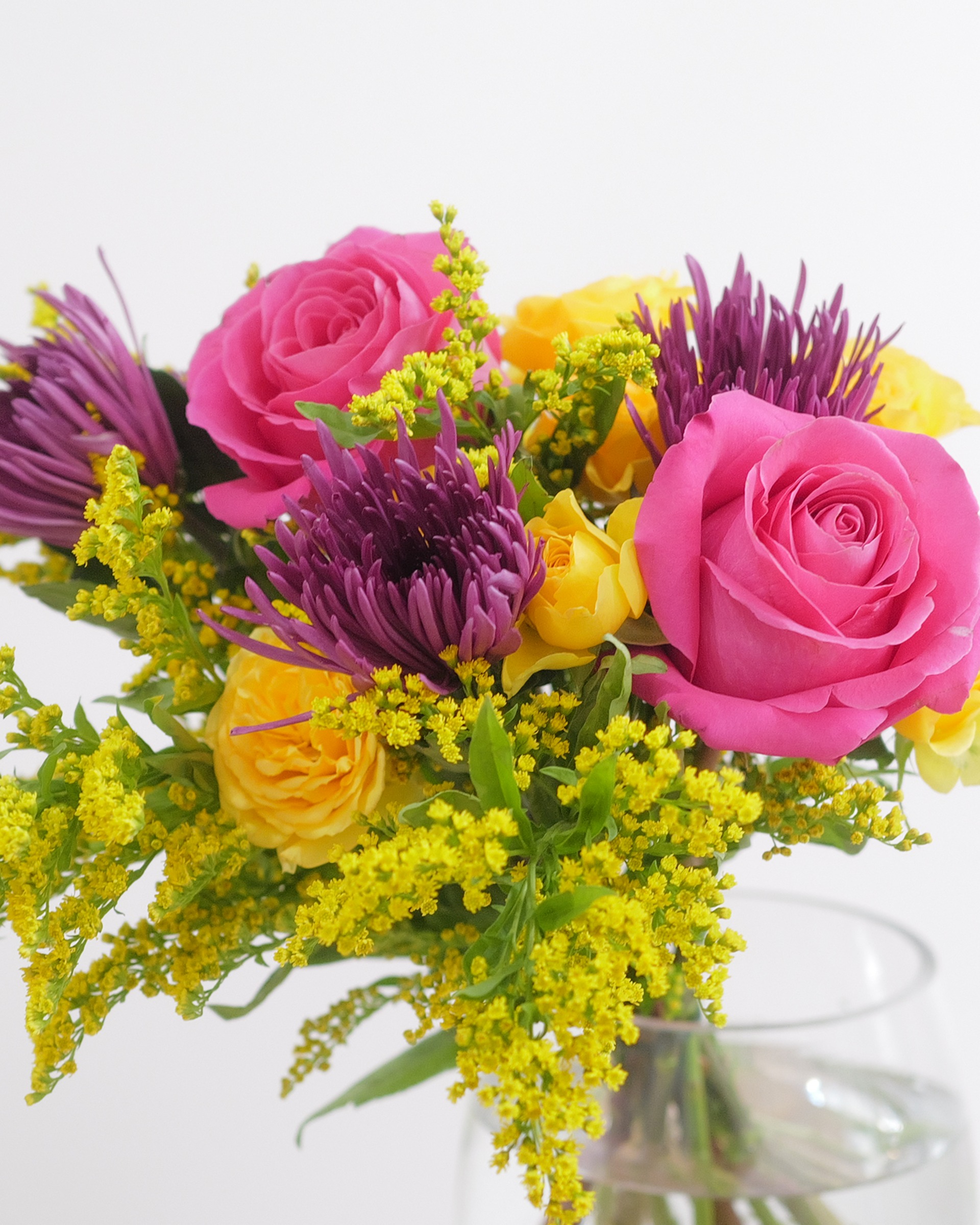 Pink roses, purple spider mums and yellow roses with sprays of bright yellow filler in a clear glass vase