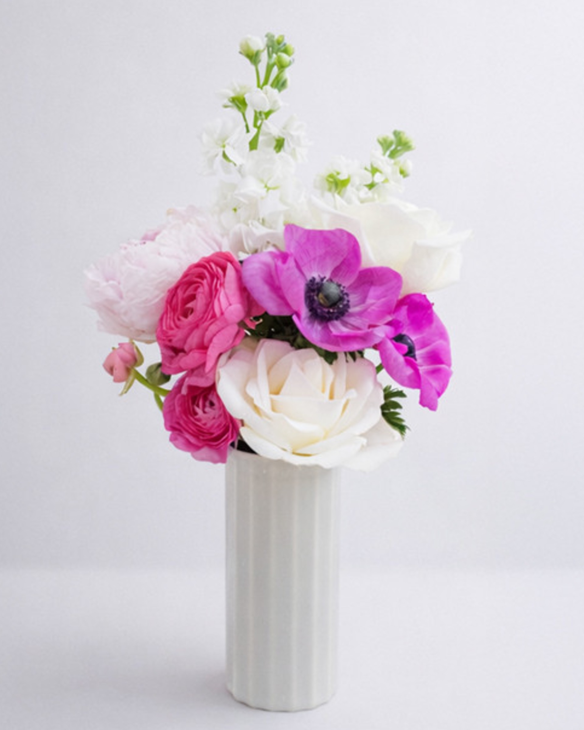 Magenta anemones, pink ranunculus, cream roses, a blush peony and white stock in a fluted white vase on a pale backdrop