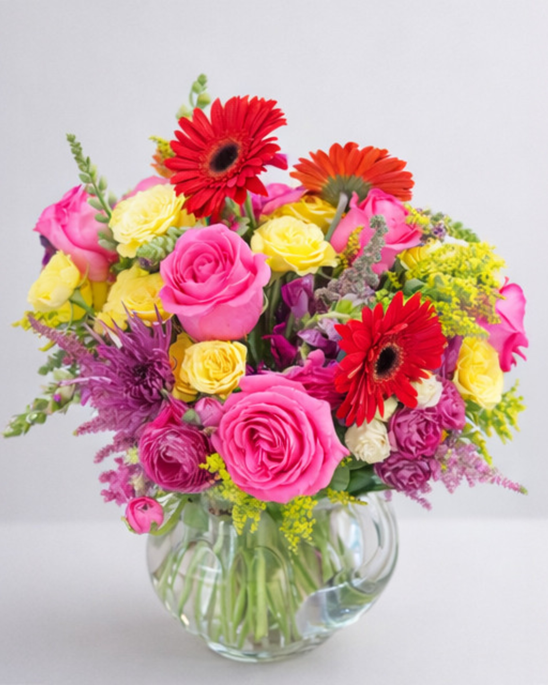 Red and orange gerberas with pink and yellow roses, purple accents and yellow solidago in a round glass vase
