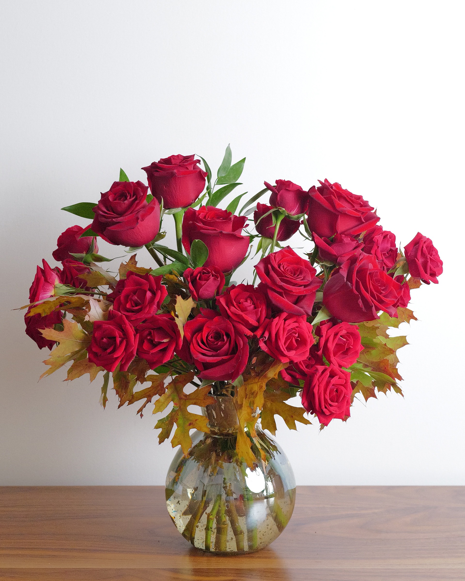 Fall into Romance, red roses and spray roses with amber autumn leaves in a round clear glass vase, seen front on