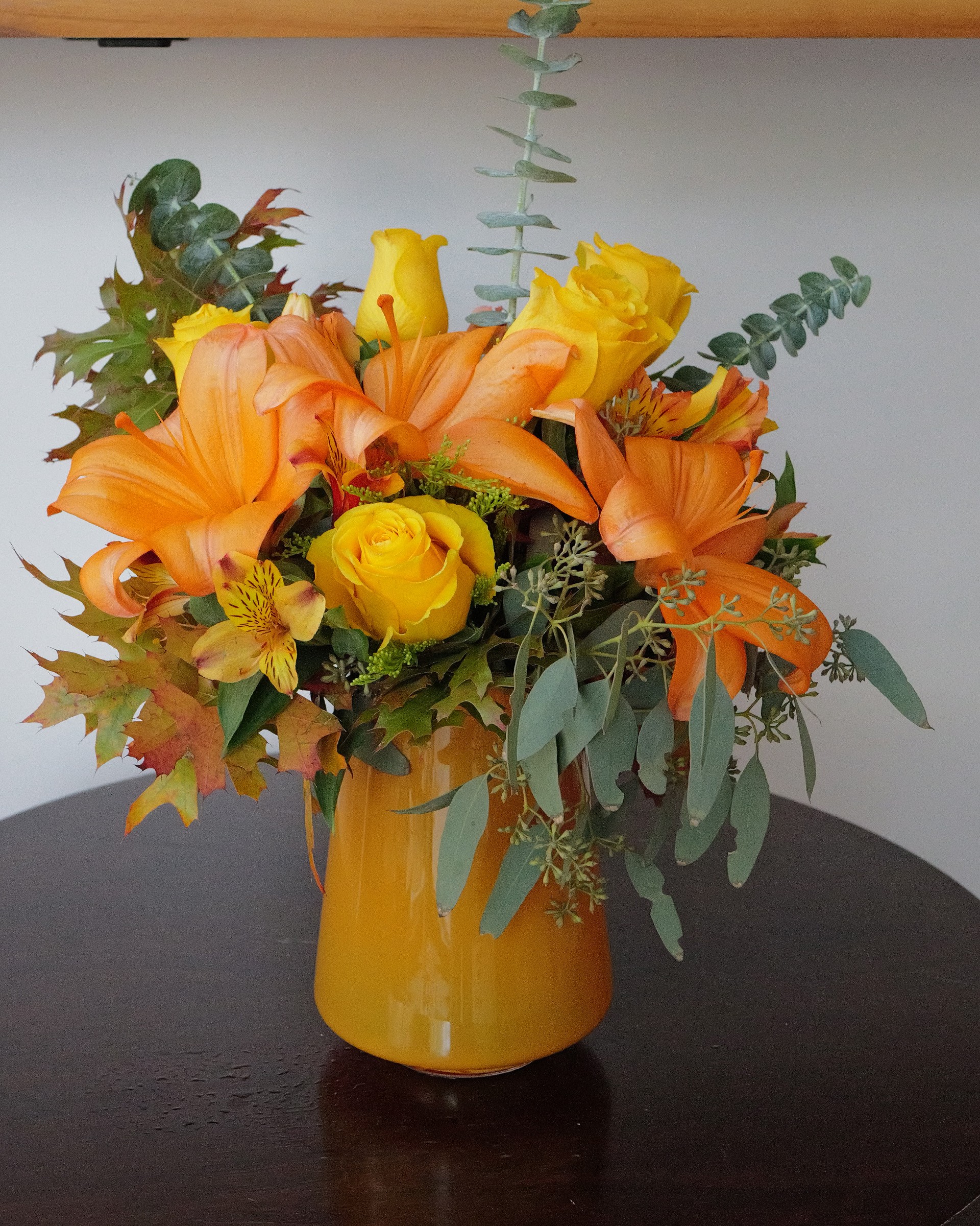 Orange lilies and yellow roses with eucalyptus and autumn leaves in an orange glass vase on a dark round table