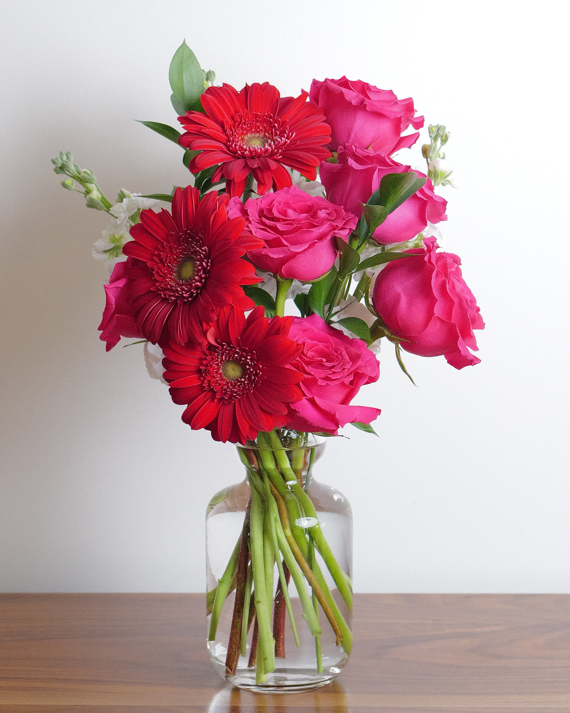 Red gerbera daisies and hot pink roses with white blooms and green leaves in a clear glass vase on a wooden table