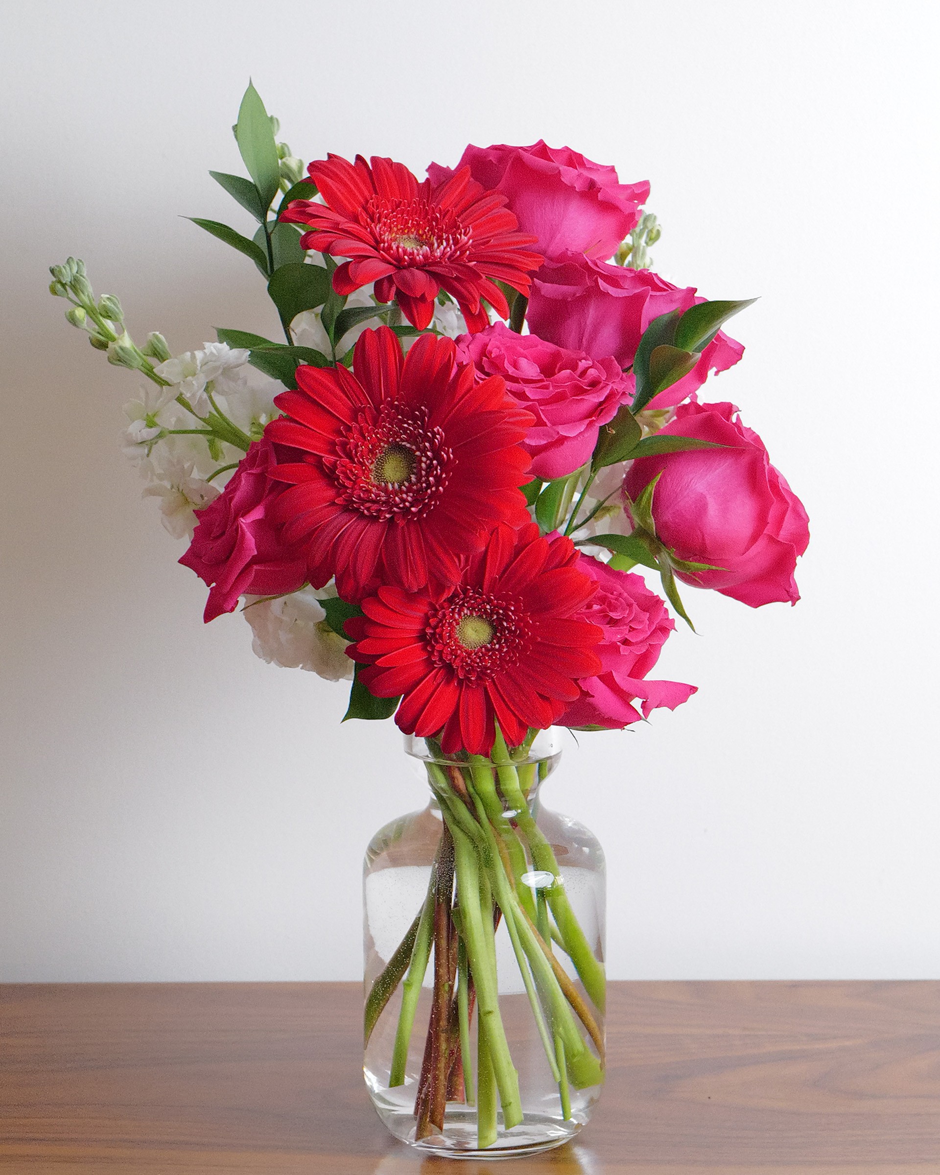 Bright red gerberas and hot pink roses with touches of white and greenery in a clear glass bottle vase on wood