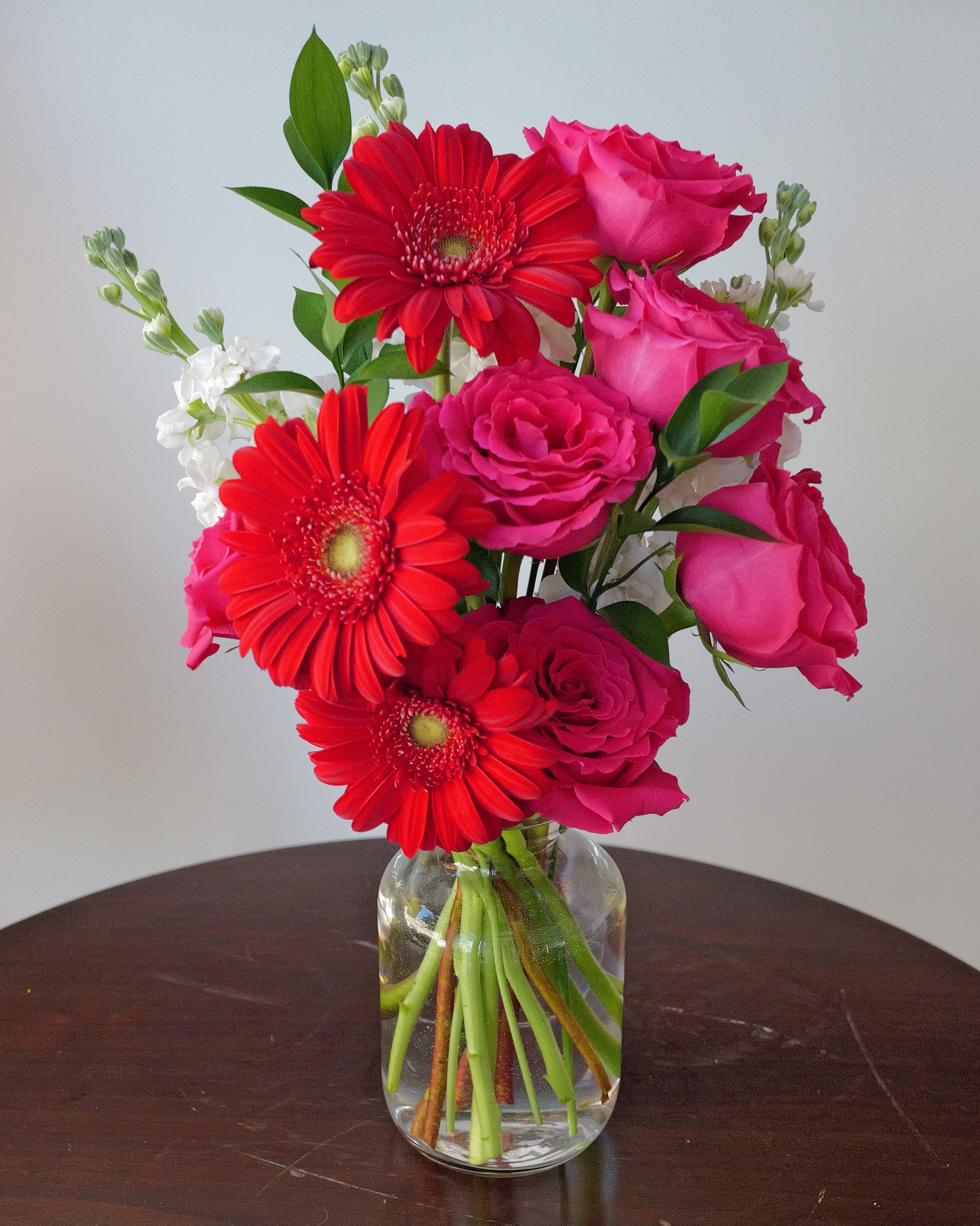 Red gerbera daisies and hot pink roses with white flowers and leaves in a clear glass vase on a dark round table