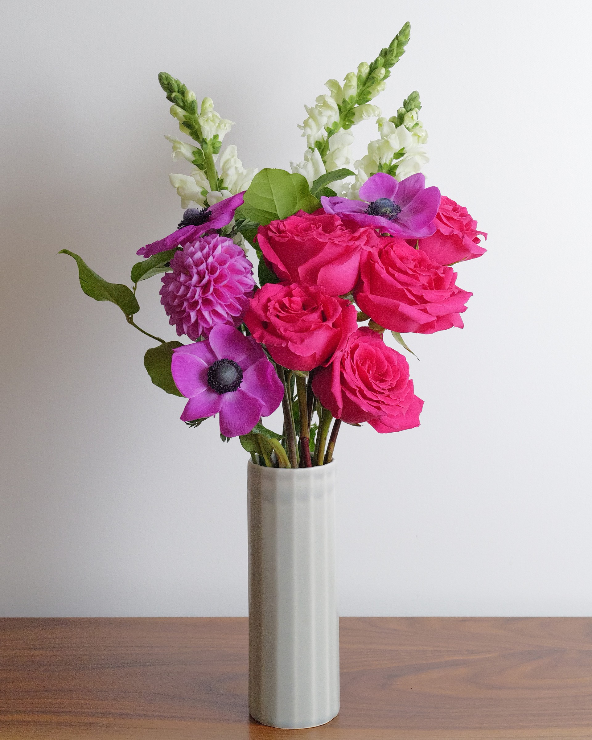 Hot pink roses, magenta anemones and white snapdragons in a tall pale ceramic vase on a wooden table.