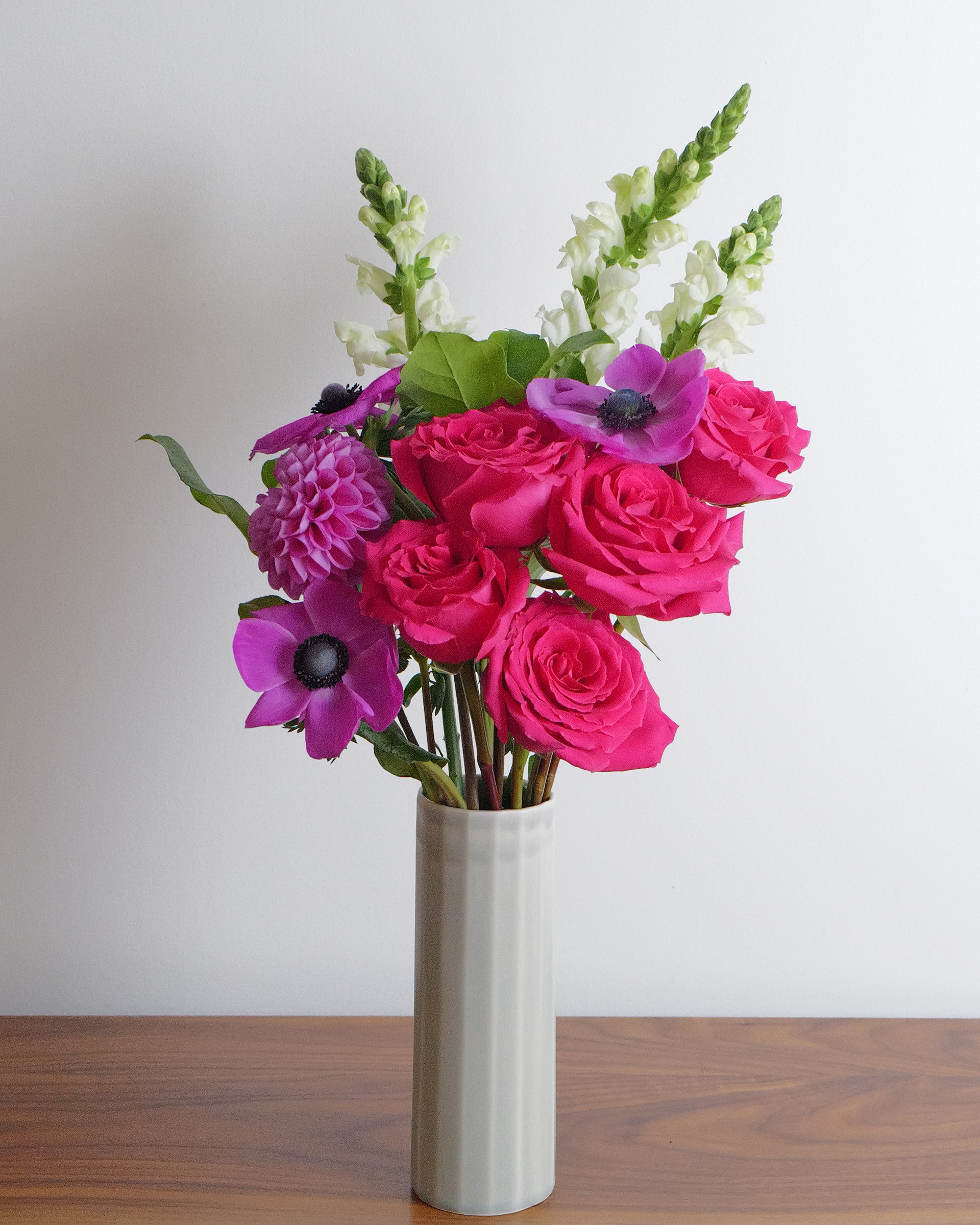 Bright pink roses and purple anemones with tall white snapdragons in a slim grey vase against a white wall