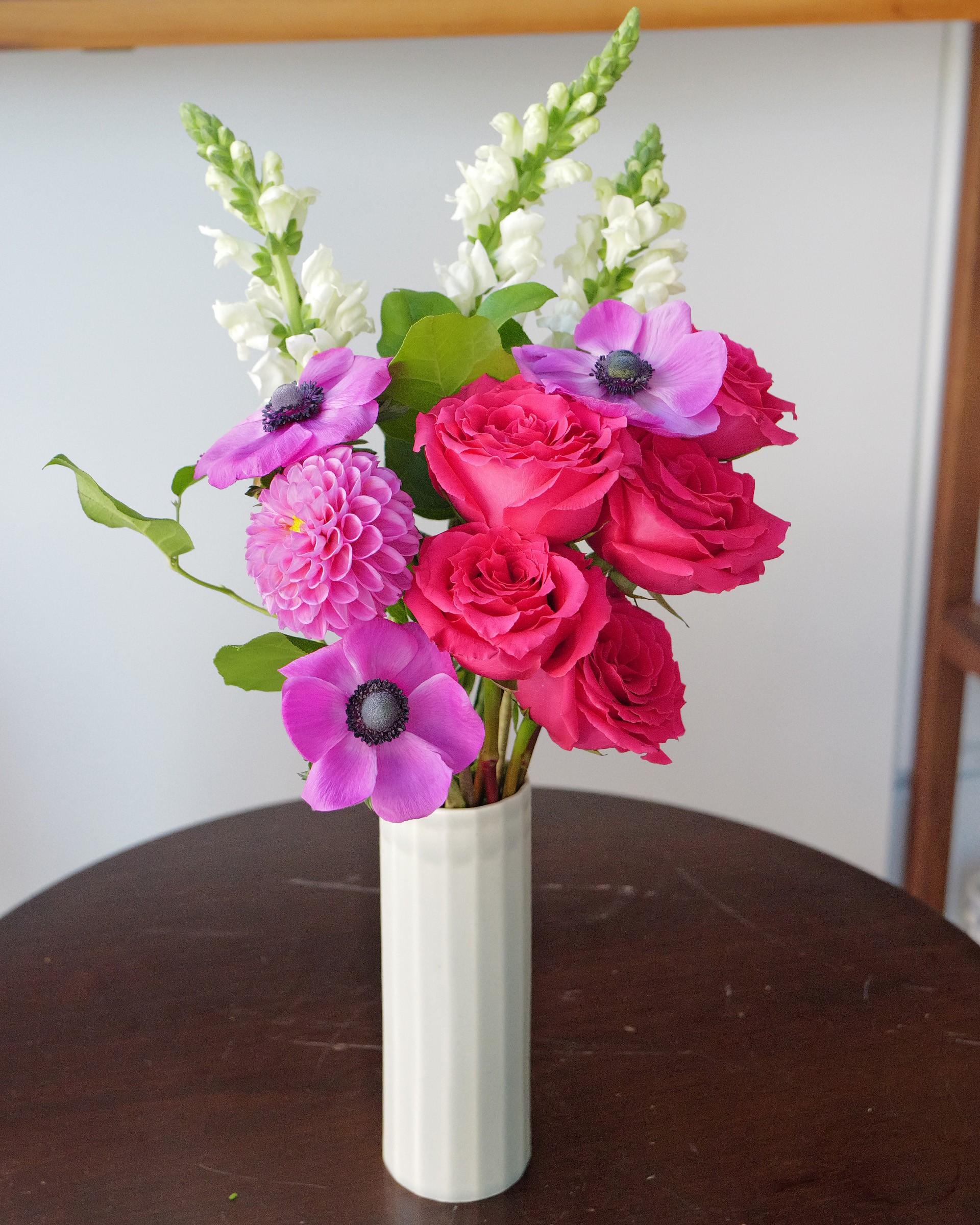 Pink roses, anemones, a pink dahlia and white snapdragons in a ribbed white vase on a dark round table