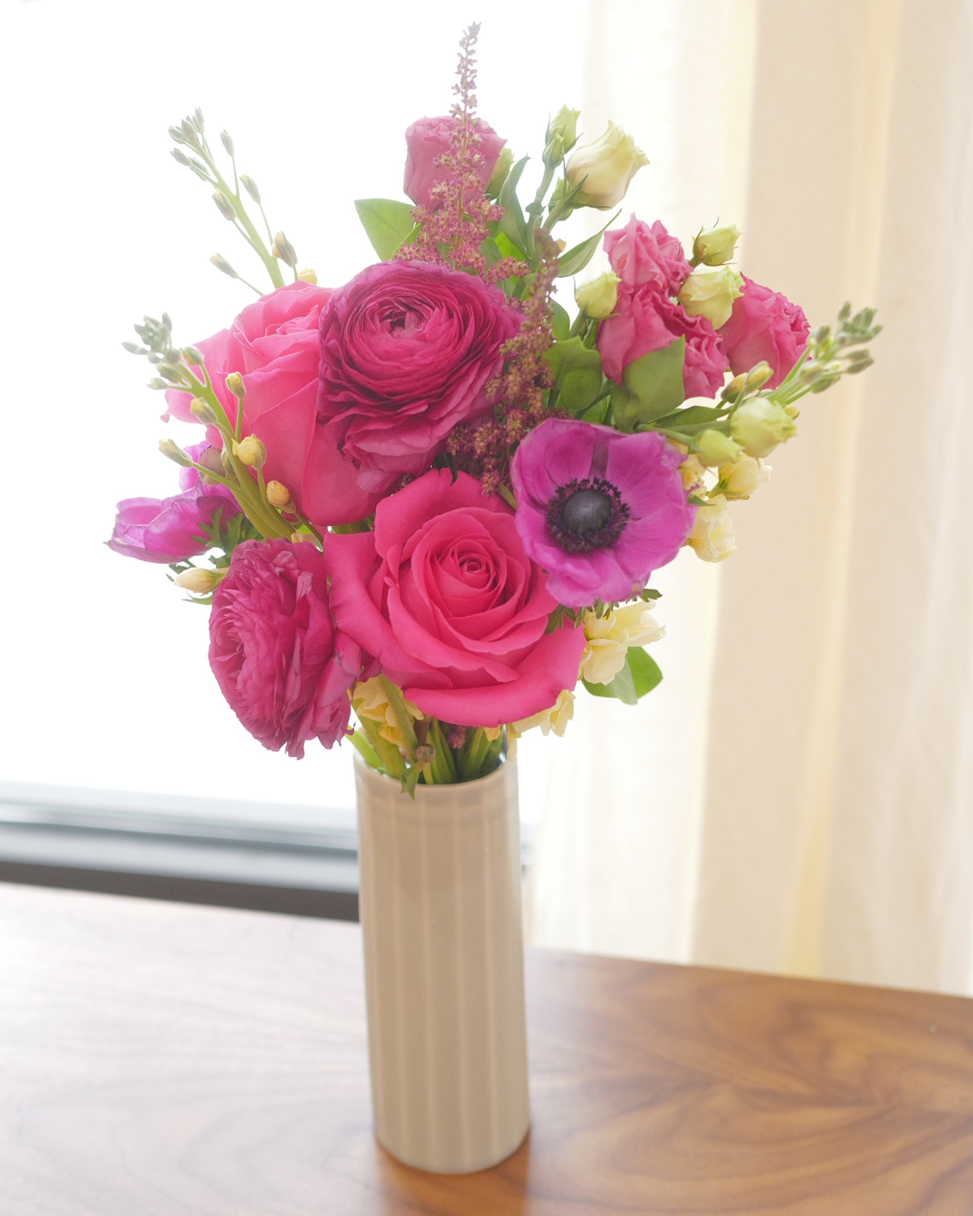 Pink roses, magenta ranunculus and an anemone with pale yellow blooms in a ribbed vase by a sunlit window
