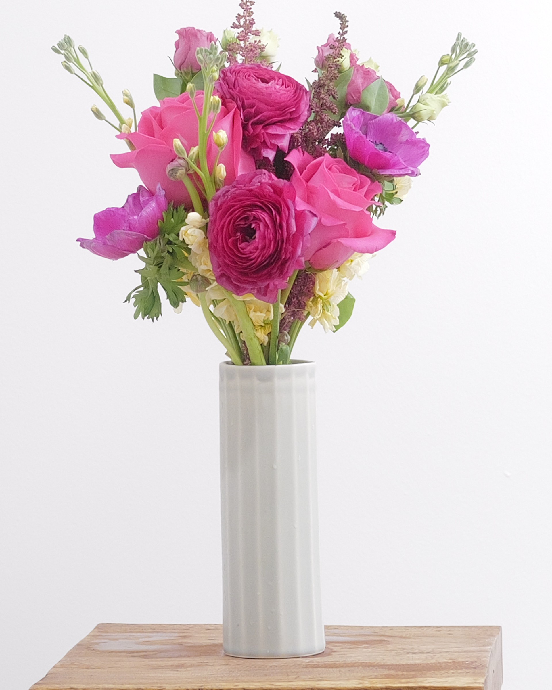 Pink roses, deep pink ranunculus and purple anemones with cream blooms in a pale grey vase on a wooden stool