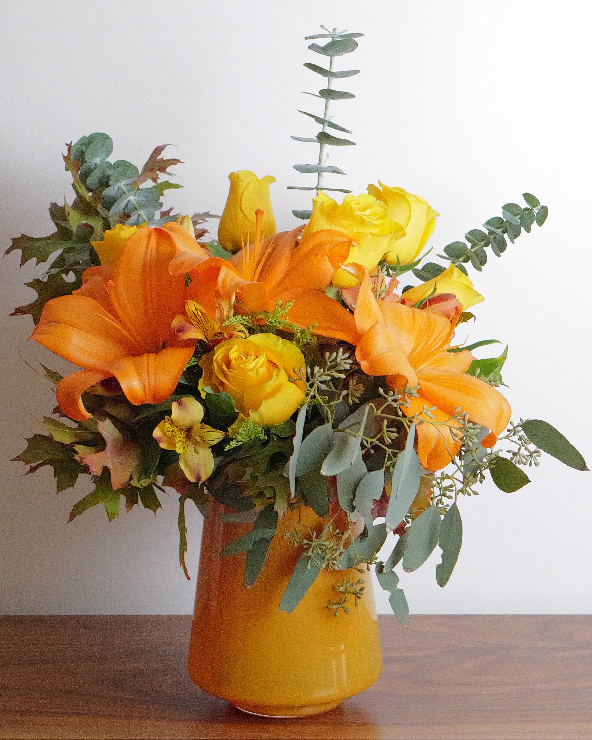 Orange lilies, yellow roses, alstroemeria and eucalyptus with autumn leaves in an orange glass vase on a wooden table
