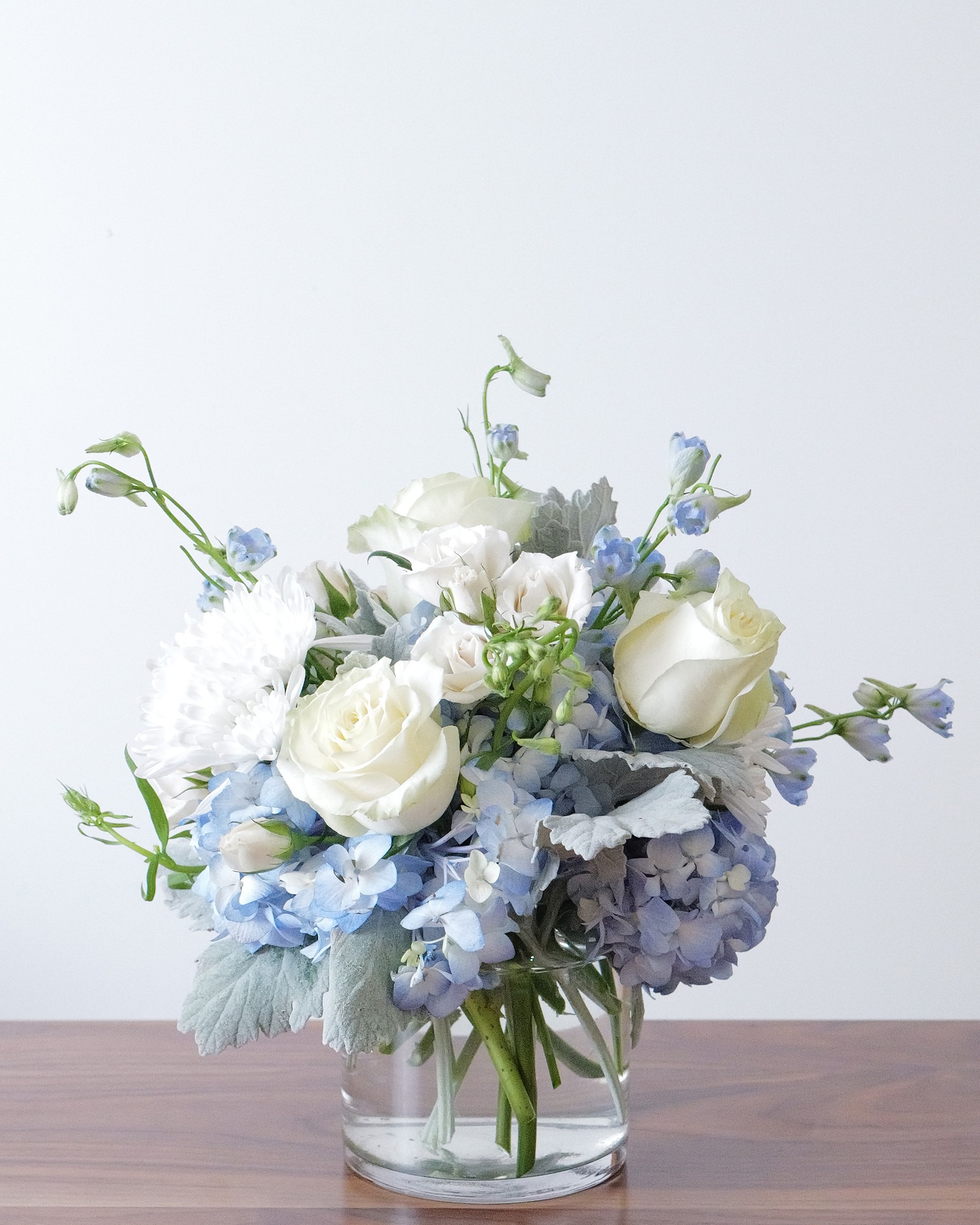 Blue hydrangea, white roses, white mums and pale blue delphinium with silvery leaves in a clear glass vase on a wood table