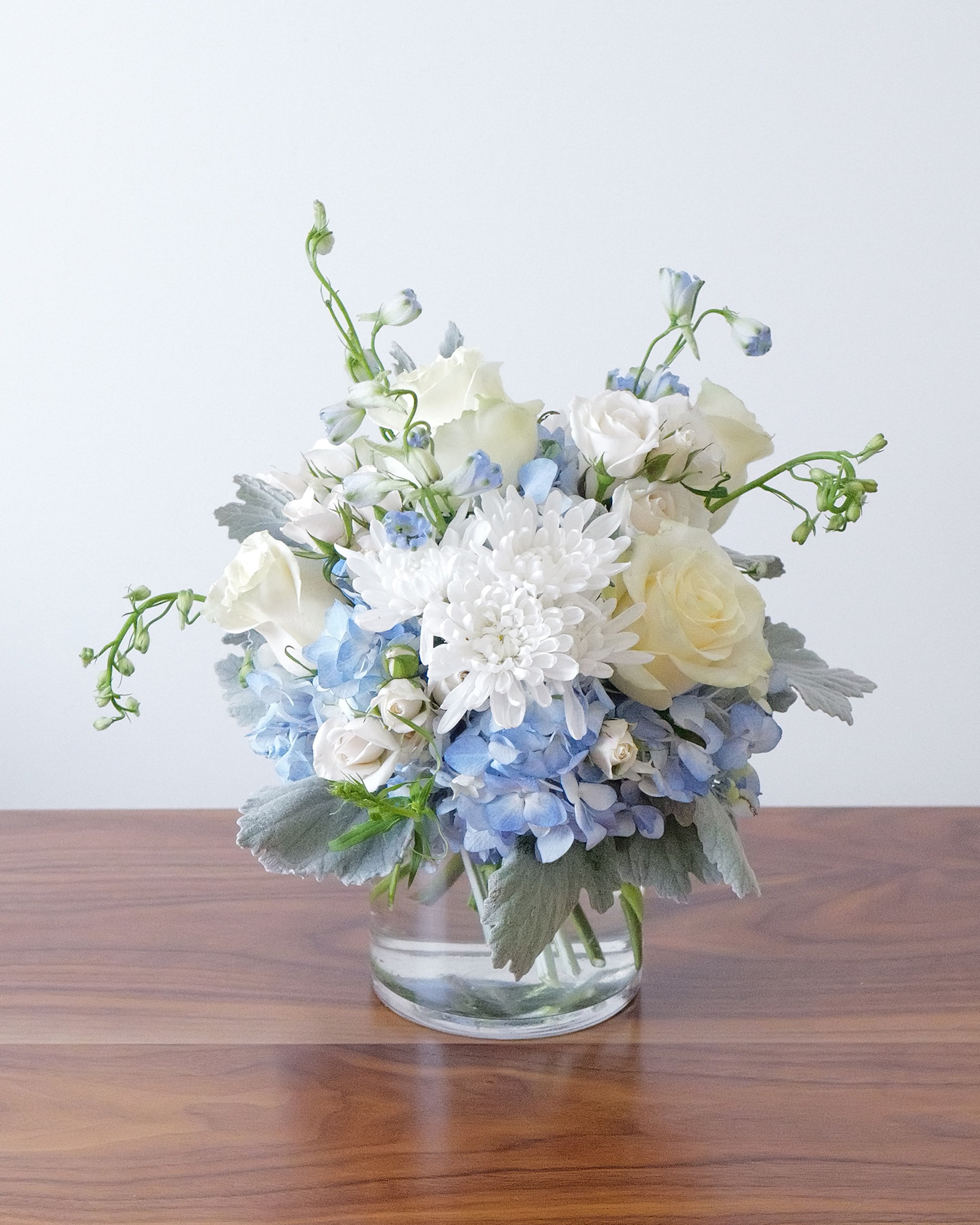 Front view of white roses and a white mum over blue hydrangea, delphinium arching out the top, in a clear glass vase