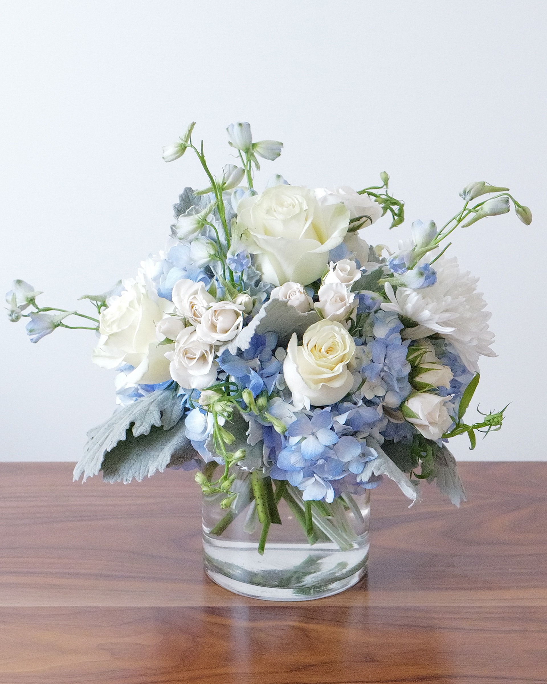 Cream spray roses, white roses and blue hydrangea with silvery leaves and blue delphinium in a clear glass cylinder vase