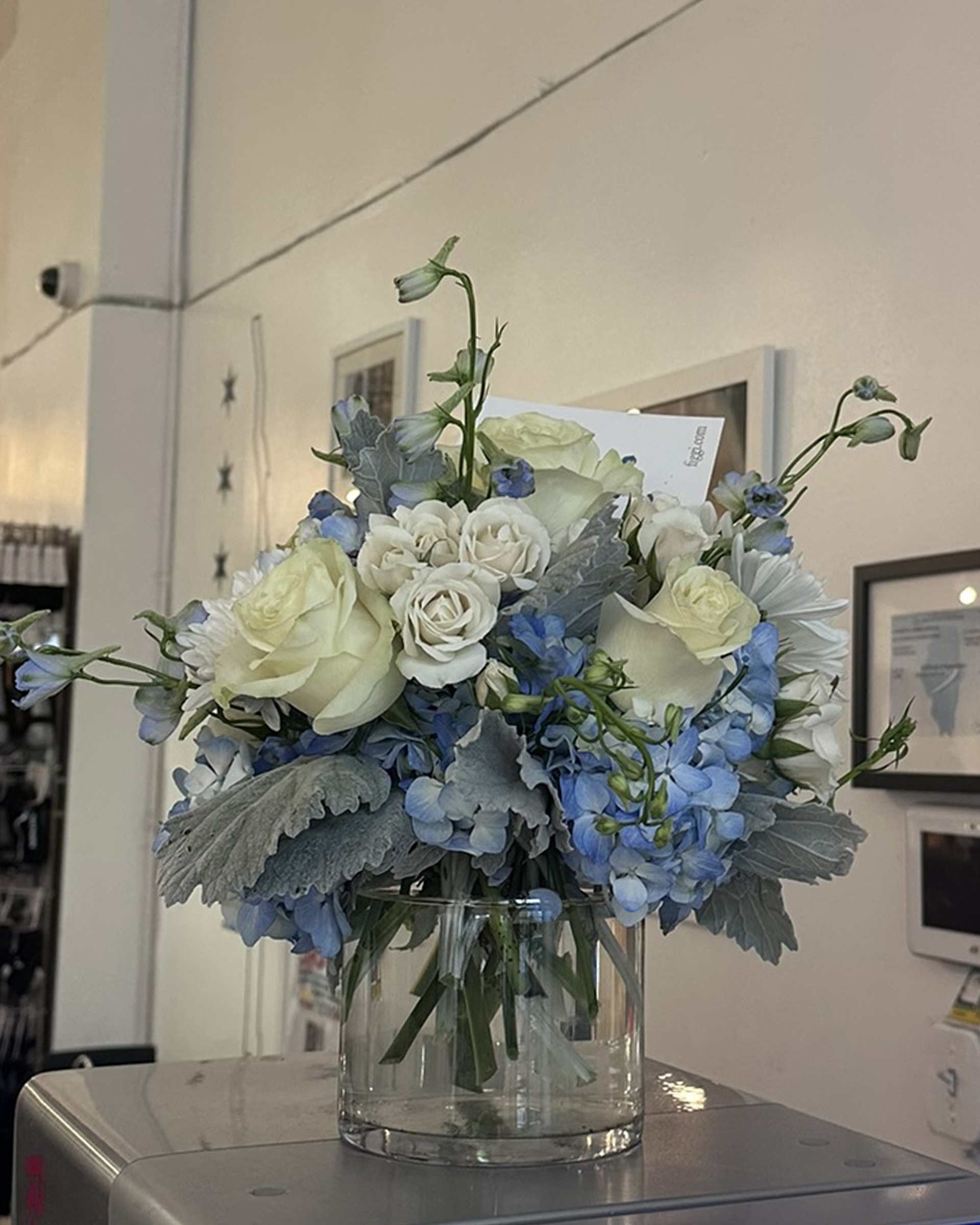 Blue and white arrangement of hydrangea, roses and delphinium in a clear glass vase with a card, on a counter in the studio